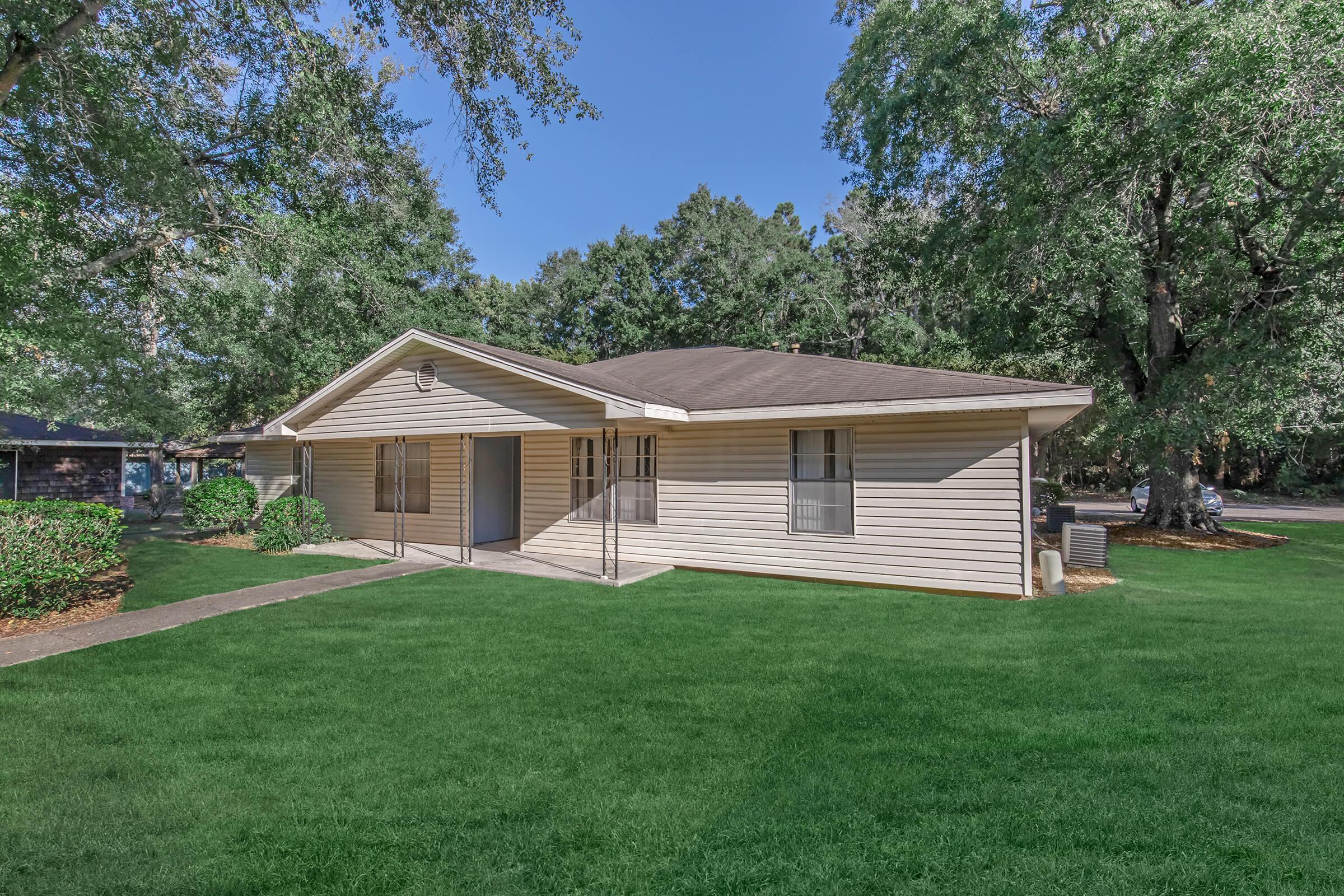 A single-story house with a tan exterior, surrounded by green lawns and trees. The house features a sloped roof, multiple windows, and a concrete walkway leading to the entrance. Bright blue sky and natural sunlight enhance the outdoor ambiance.