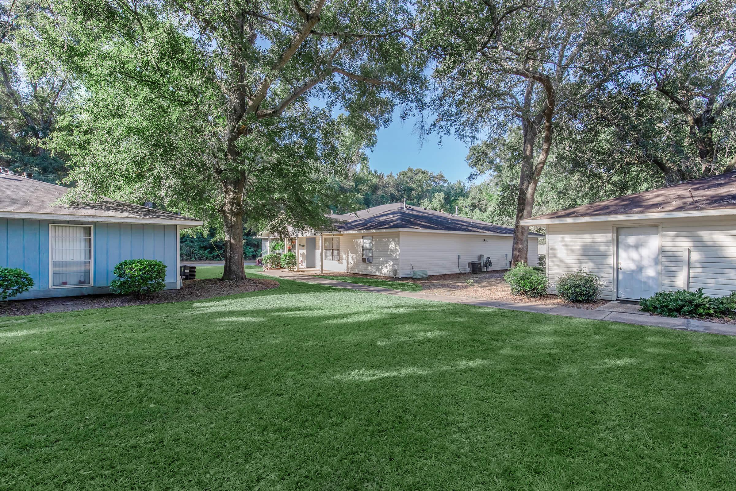 A backyard scene featuring two houses, one in blue and the other in light beige, surrounded by lush green grass and trees. The beige house has a porch visible, while the area is well-maintained, creating a tranquil and inviting outdoor space. Sunlight filters through the trees, enhancing the serene atmosphere.