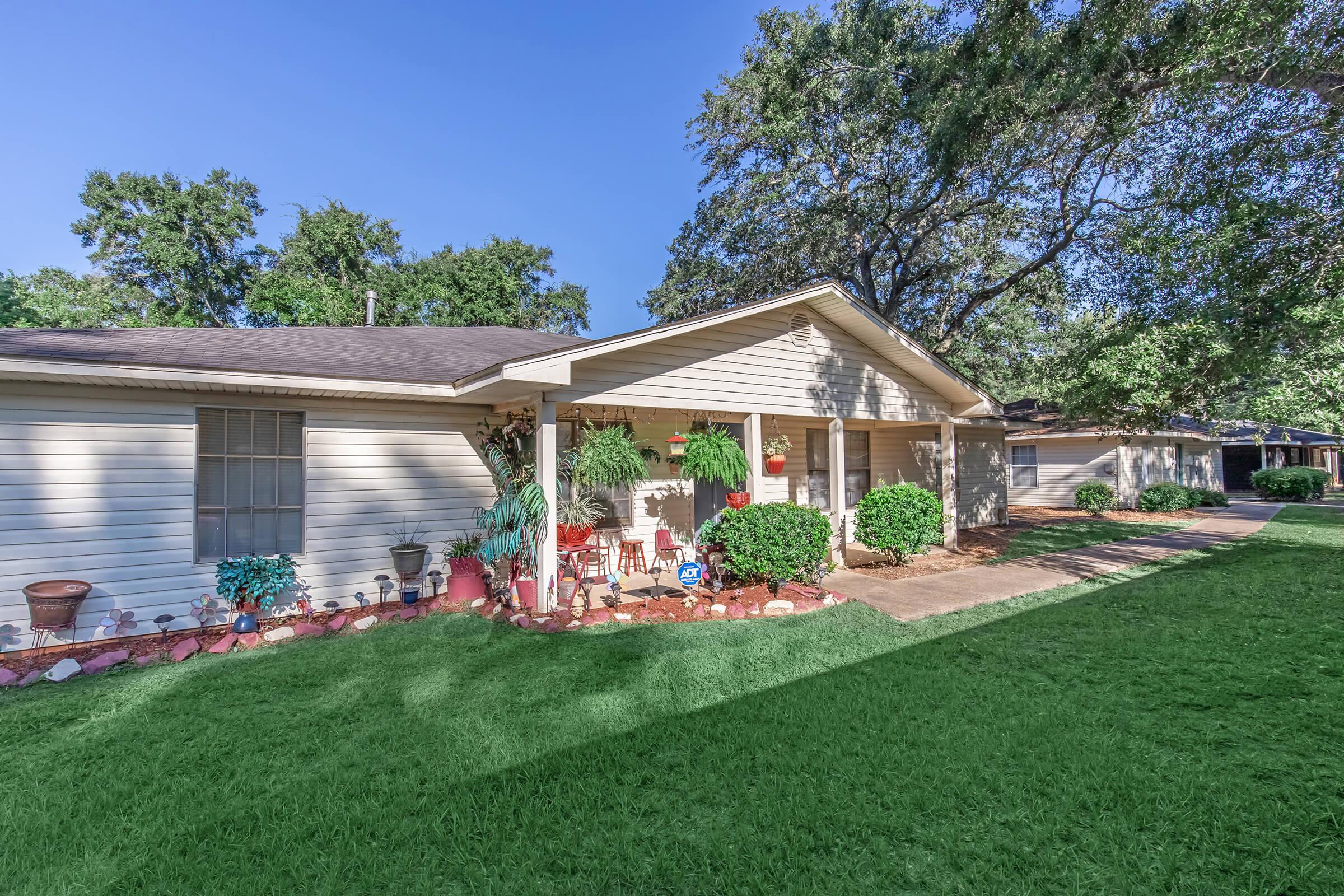 A charming single-story house with a covered porch adorned with potted plants, welcoming decorations, and a manicured lawn. The facade features light-colored siding and a clear blue sky overhead, surrounded by trees. The driveway leads towards a nearby garage, creating an inviting and well-kept appearance.