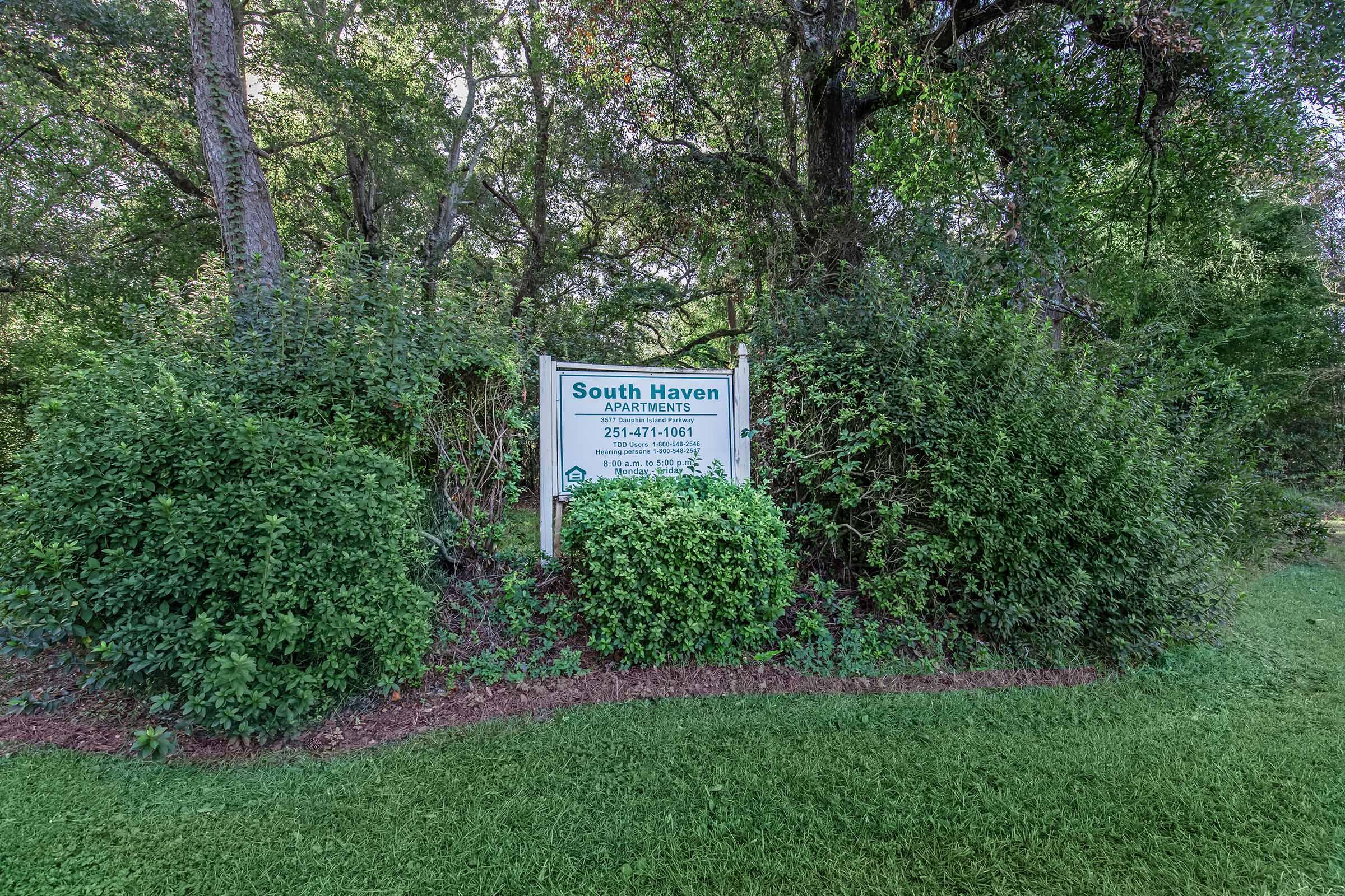 Sign for South Haven Apartments surrounded by shrubs and trees, featuring contact information and a welcoming message. The sign is positioned in a green, landscaped area, emphasizing the apartment complex's natural surroundings.