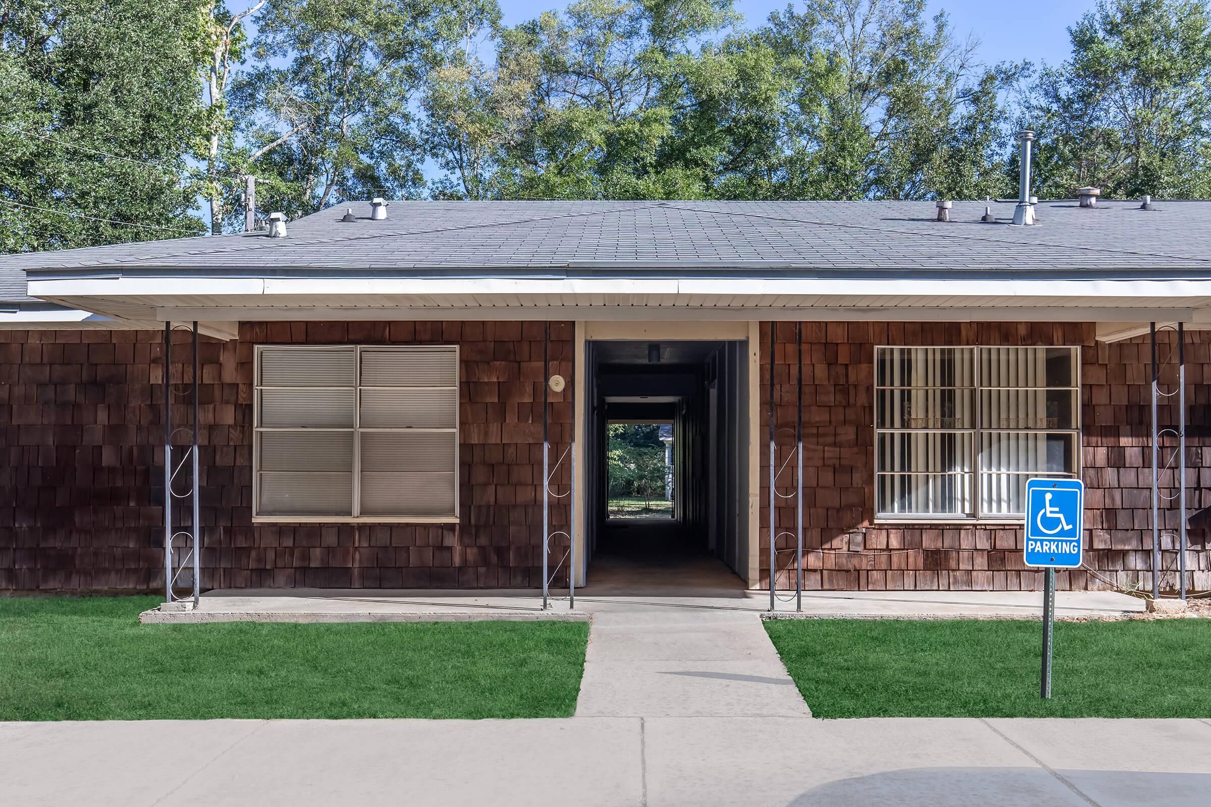 A single-story building with a wooden exterior and large windows. An accessible parking sign is visible in front, indicating designated parking for individuals with disabilities. The entrance has a concrete path leading to a shaded area in the background surrounded by trees.