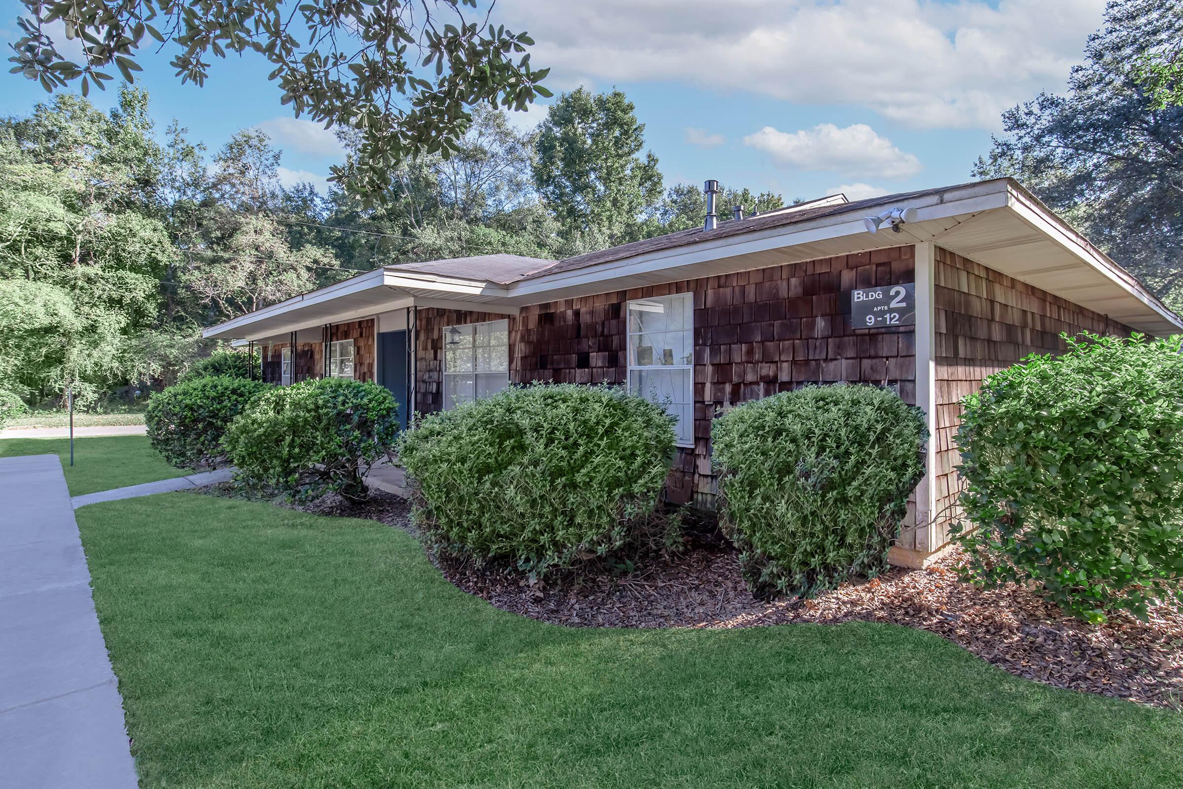 A single-story residential building with wooden shingles and two front windows, surrounded by neatly trimmed bushes and a grassy lawn. The path leading to the entrance is made of concrete, and trees are visible in the background under a partly cloudy sky.