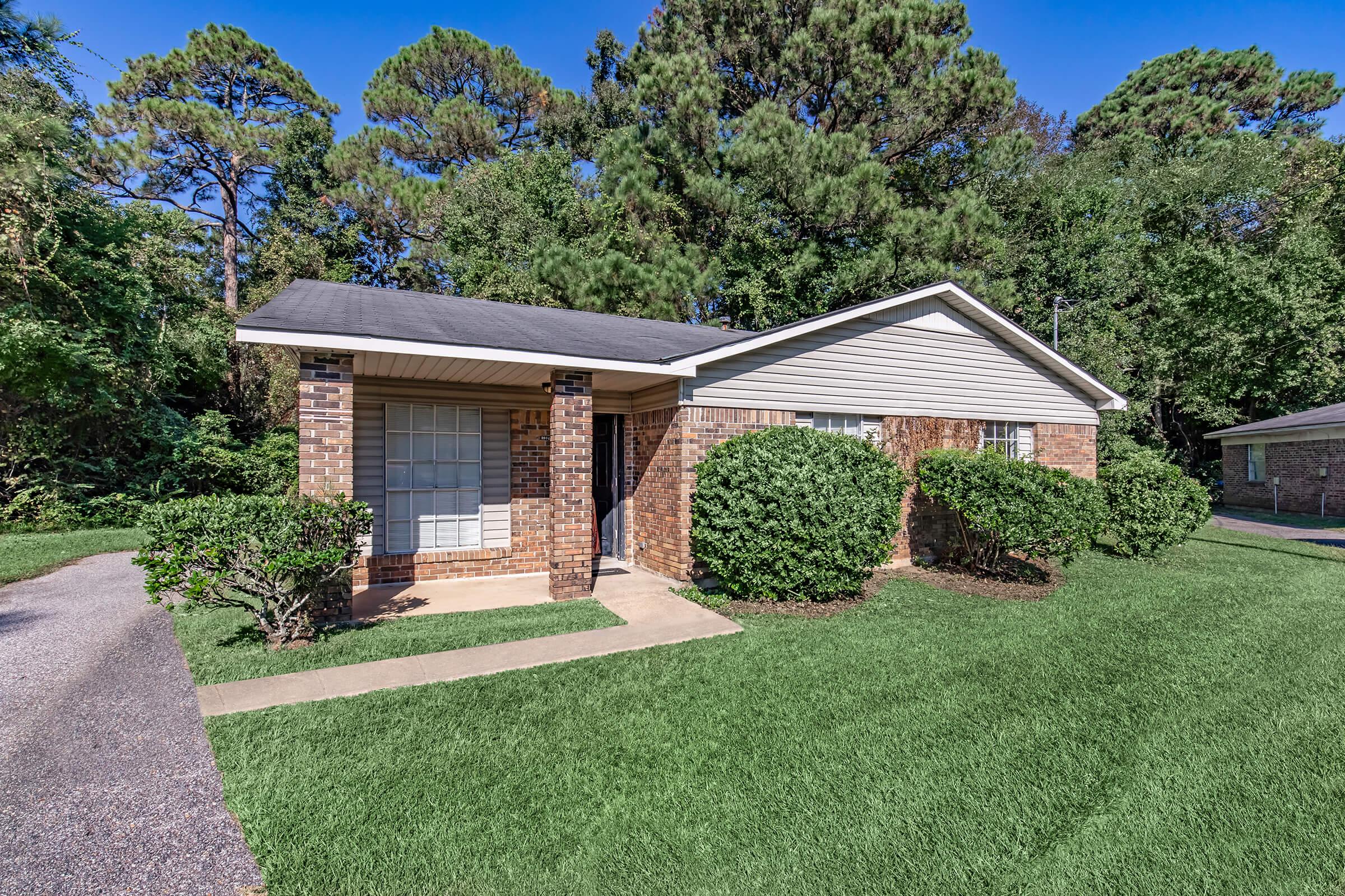 Single-story brick house with a gabled roof, surrounded by well-maintained shrubs and grass. The front porch has a small walkway leading to the entrance. Lush green trees and vegetation in the background provide a natural setting, with clear blue skies above.