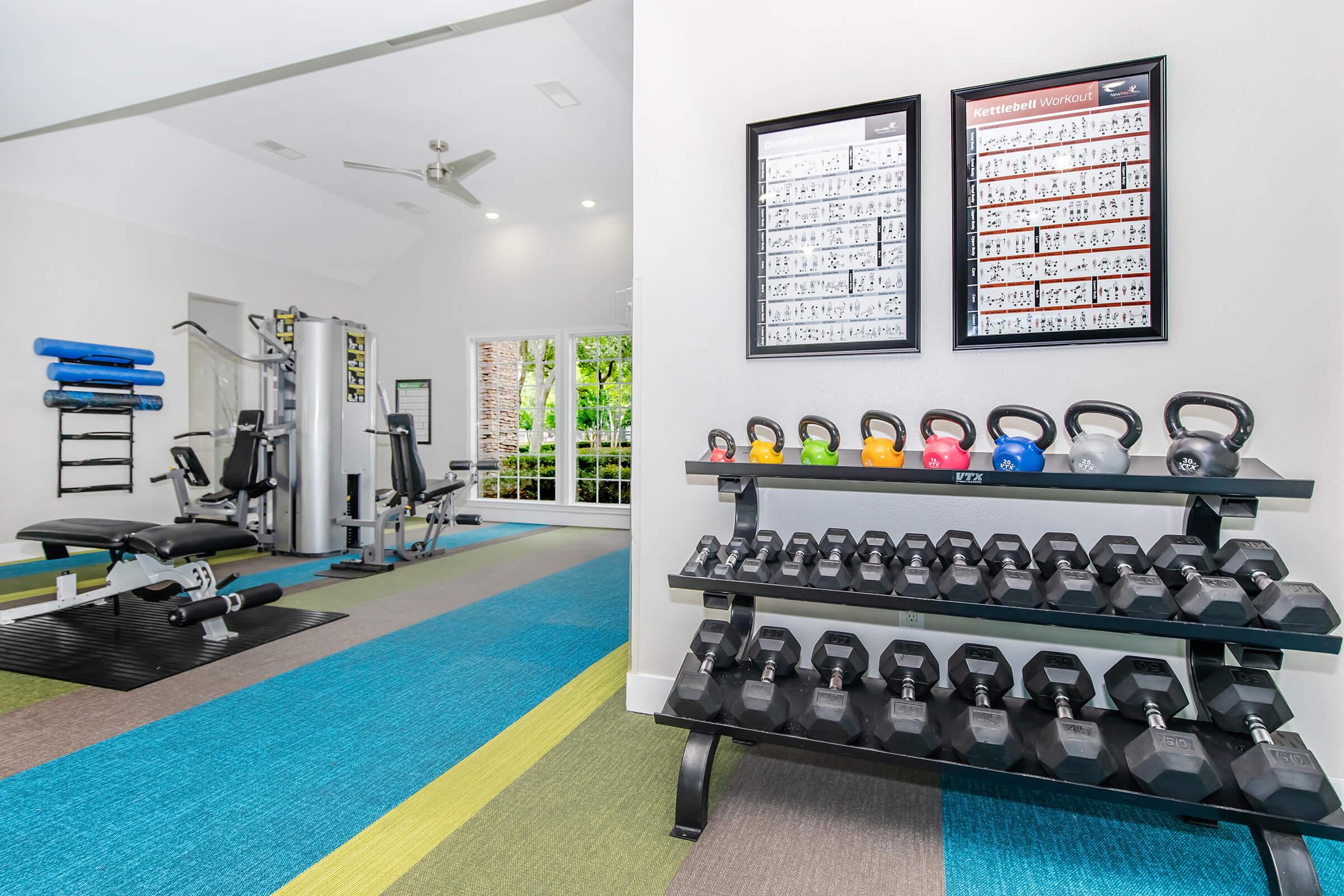 A well-lit home gym featuring a variety of workout equipment. There is a rack of colorful kettlebells and dumbbells, a multi-station weight machine, exercise mats, and instructional posters on the wall. The flooring has a mix of vibrant colors, and large windows allow natural light to brighten the space.