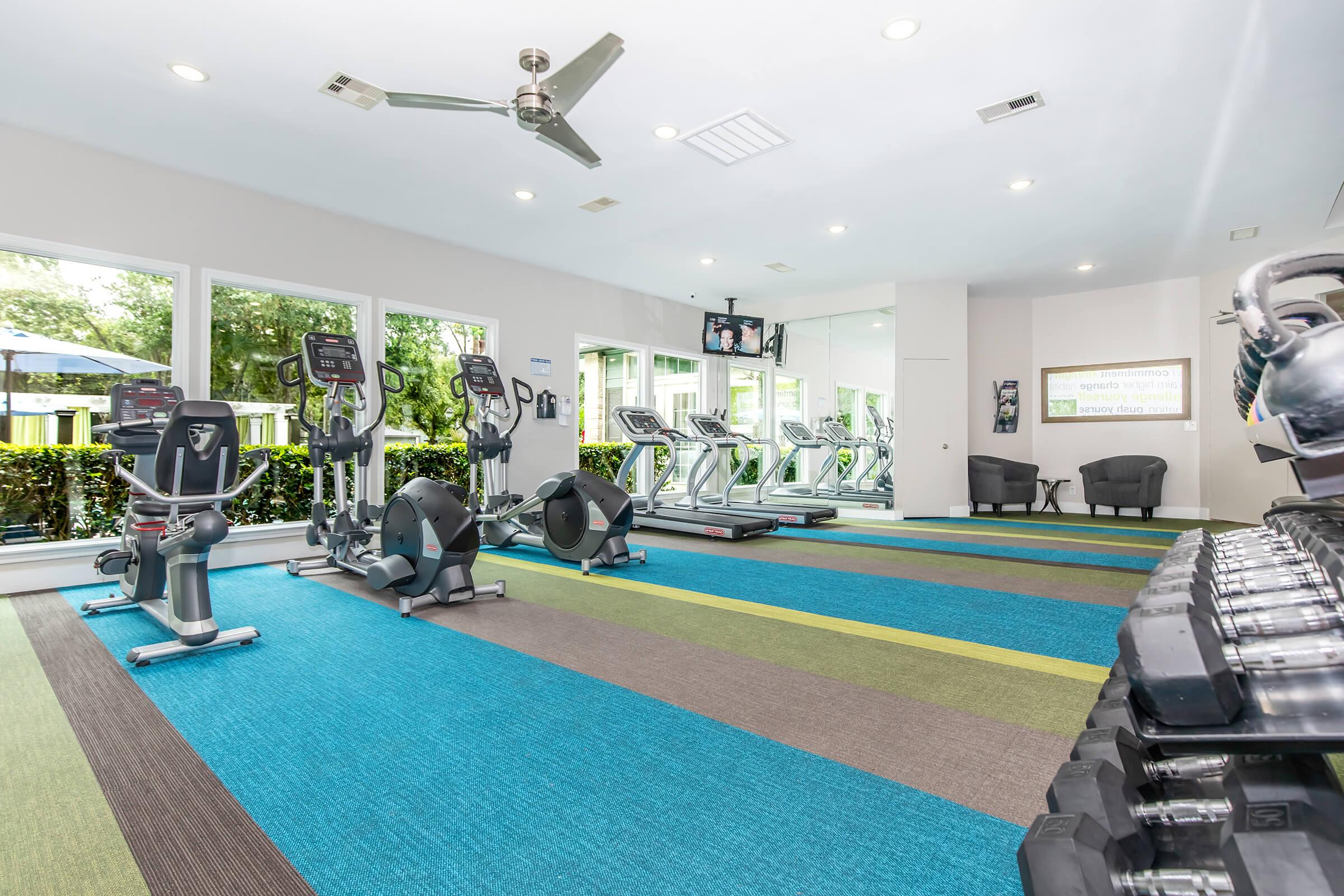 Fitness room featuring various exercise equipment, including treadmills, ellipticals, and stationary bikes. The space has colorful carpeting, large windows allowing natural light, and modern decor, creating an inviting atmosphere for workouts.