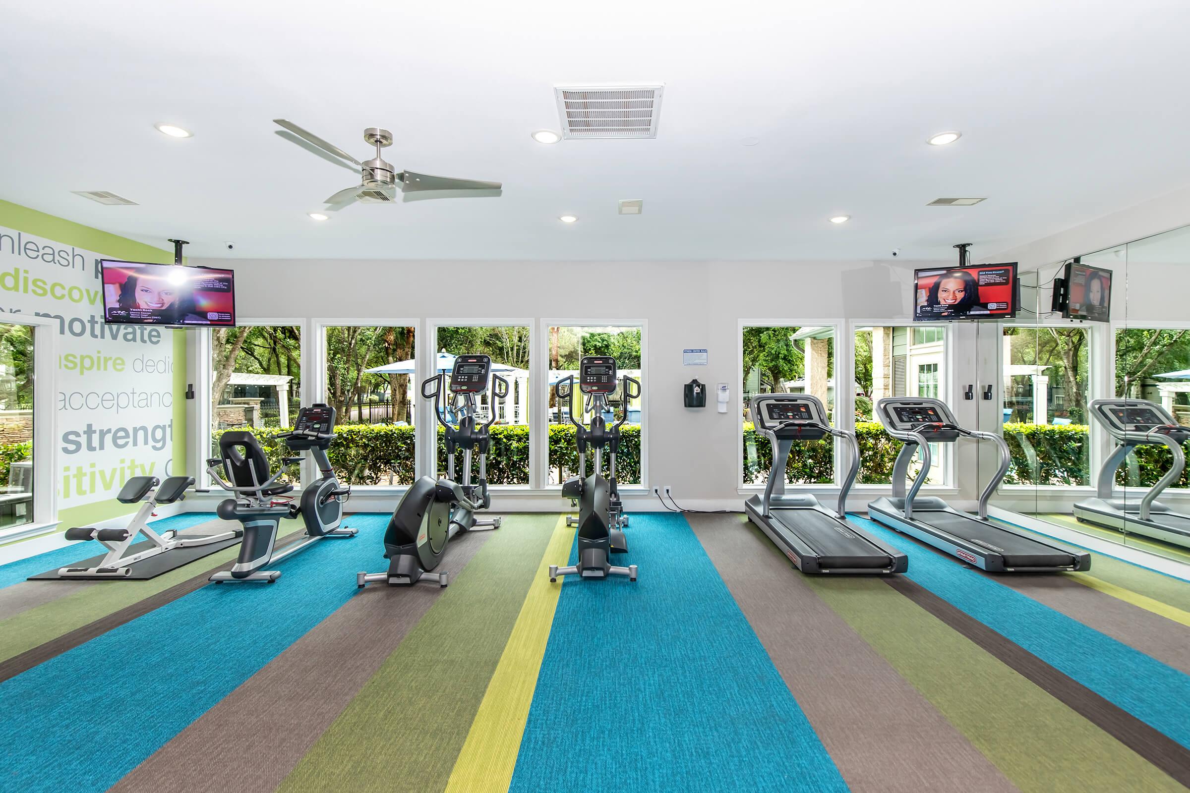 A modern gym with exercise equipment, including treadmills and stationary bikes, arranged neatly on colorful carpet. Large windows allow natural light, and motivational text is displayed on a wall. Two televisions mounted on the wall are visible, enhancing the workout environment.