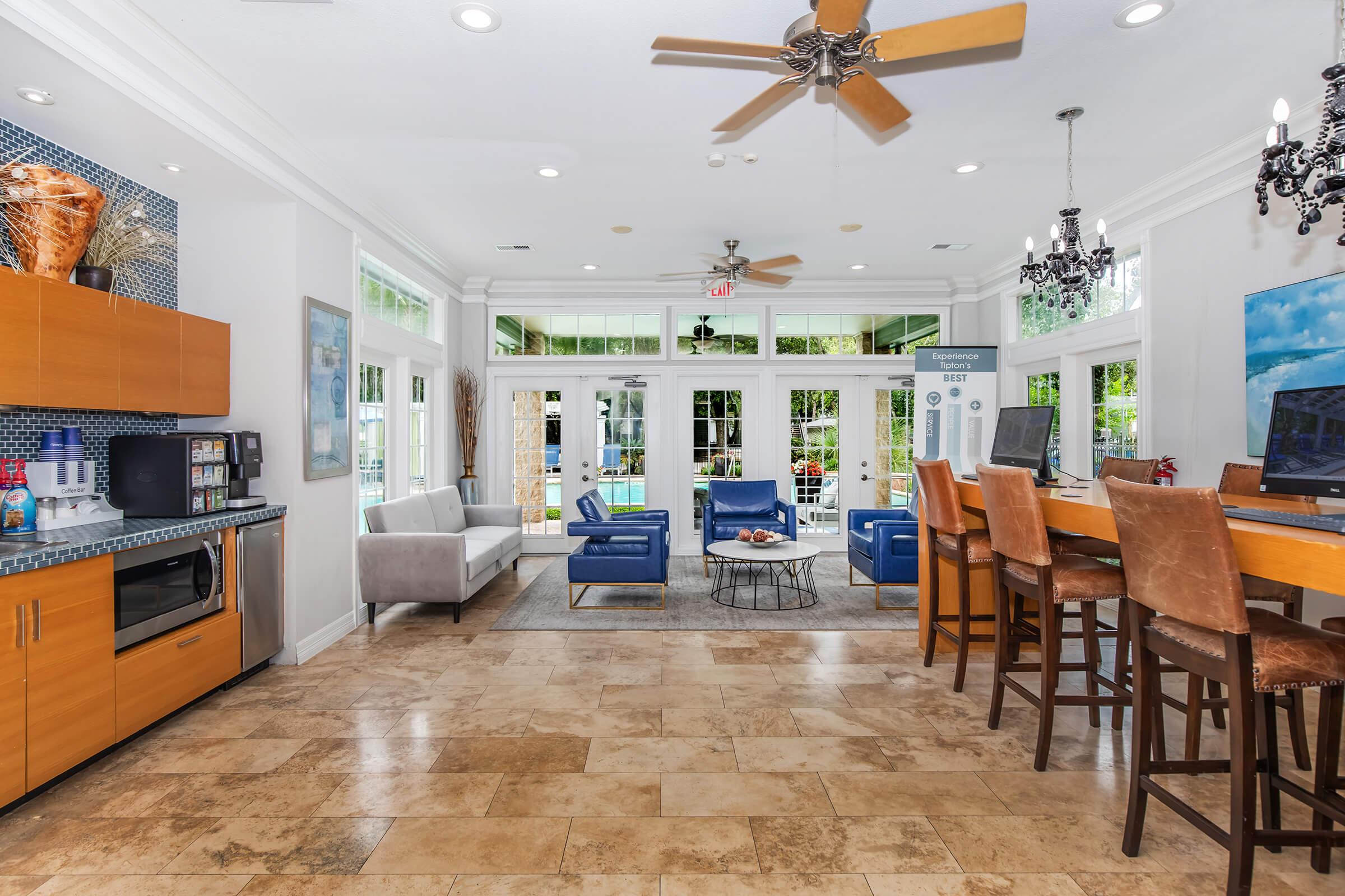 Interior view of a modern lounge area featuring a combination of seating options. Light-colored walls and a tiled floor complement wooden furniture, including a kitchenette with a microwave and beverages. Large glass doors lead to an outdoor pool area, with ample natural light from multiple windows and overhead lighting fixtures.