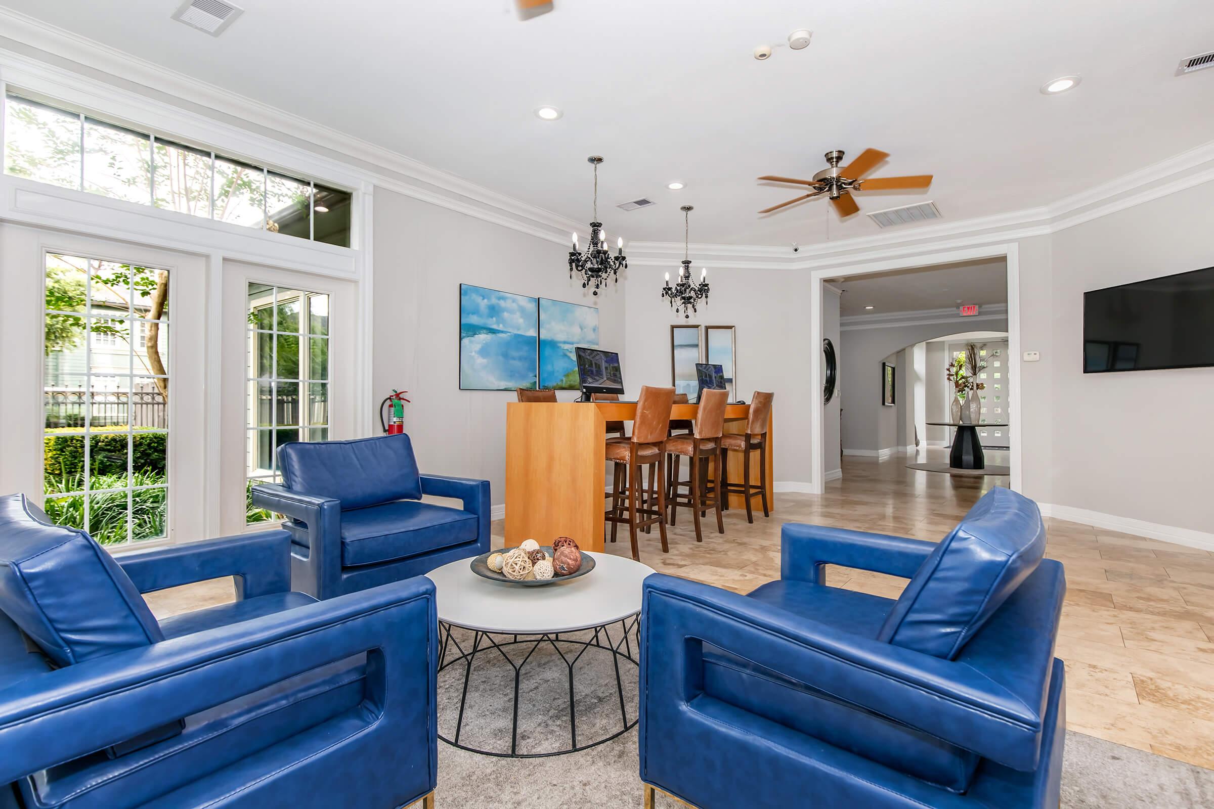 A modern living space featuring two blue leather chairs, a round coffee table with decorative items, and a view of a dining area with wooden chairs at a bar. Large windows bring in natural light, and there are decorative elements like chandeliers and wall art. A lush outdoor space is visible through the windows.