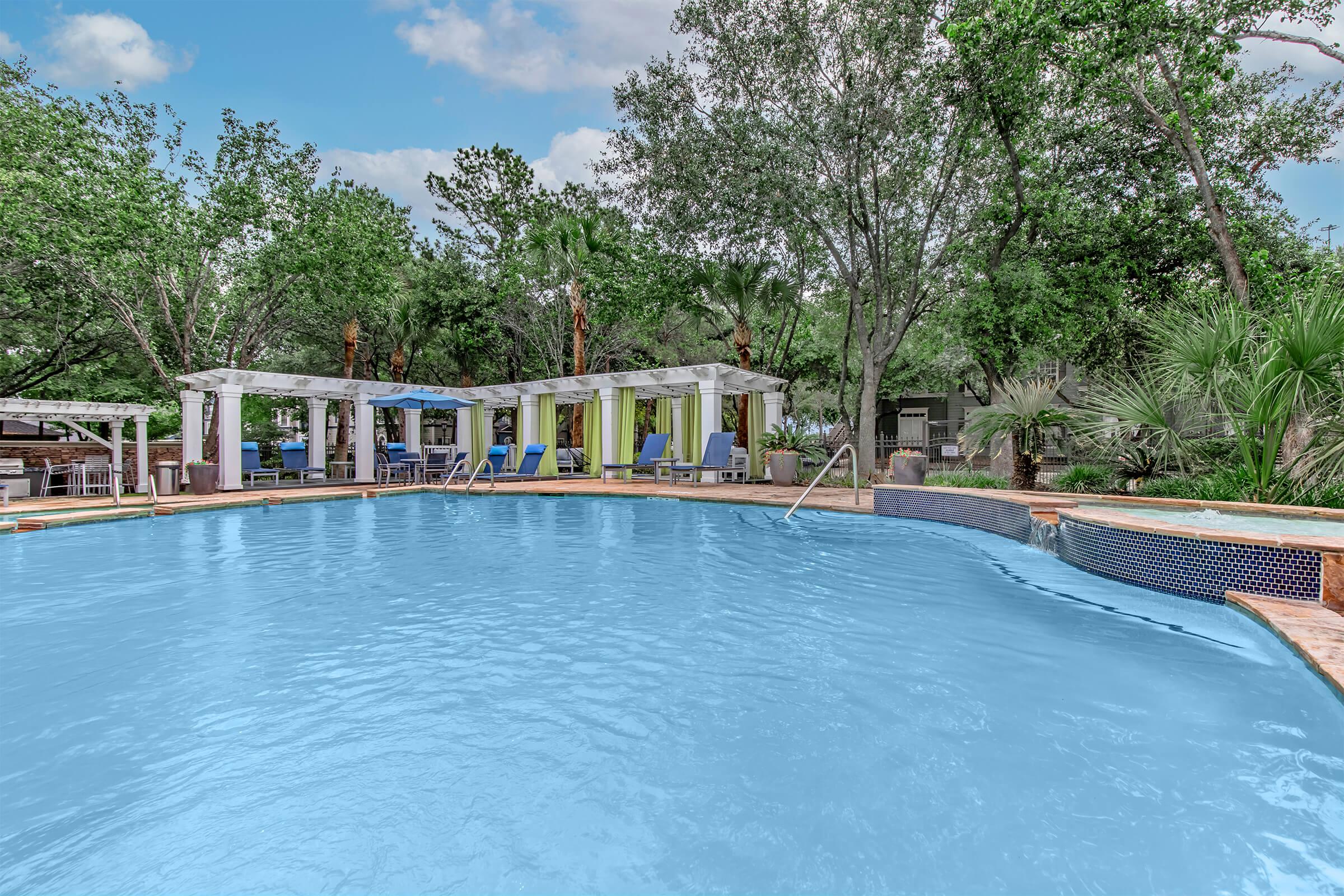 A clear blue swimming pool surrounded by lush greenery. Poolside lounges are visible, along with shaded areas featuring white curtains and colorful seating. Trees and tropical plants provide a serene atmosphere, creating a relaxing outdoor retreat.