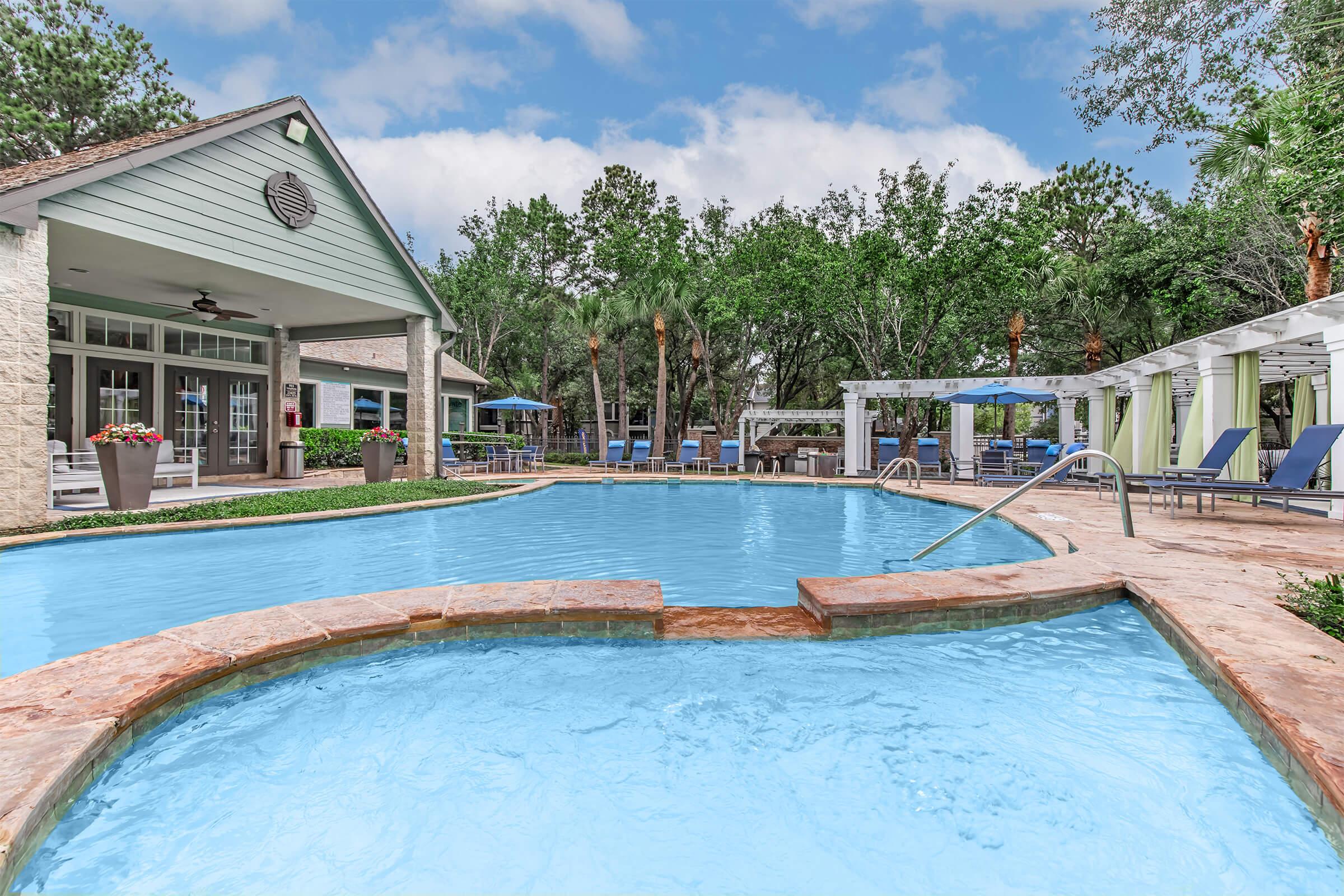 A landscaped outdoor pool with clear blue water, curving edges, and surrounding lounge chairs. In the background, a spacious building with large windows, potted flowers, and umbrellas adds a welcoming atmosphere. Lush green trees and a cloudy sky create a serene, relaxing environment.