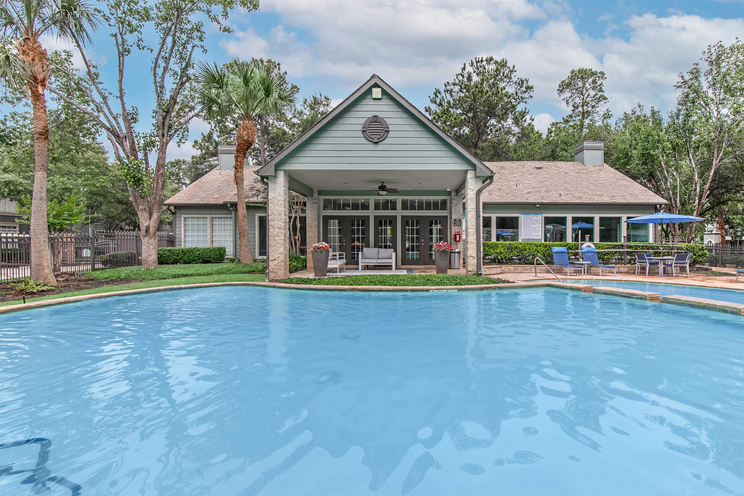 A scenic view of a swimming pool in the foreground, with a well-maintained clubhouse in the background surrounded by lush trees and palm trees. The clubhouse features large windows and an overhanging roof, with lounge chairs and tables set up nearby, ideal for relaxation and outdoor gatherings.
