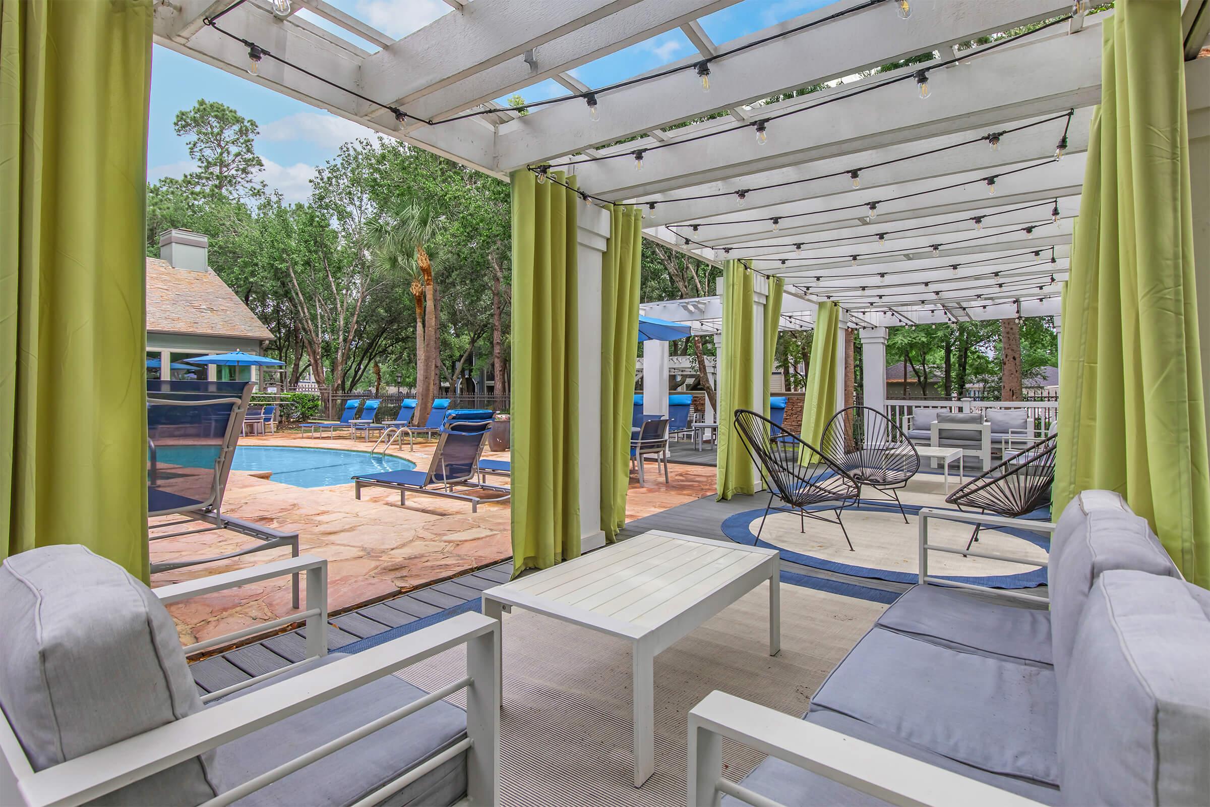 A modern outdoor lounge area with stylish gray furniture and green drapes, overlooking a swimming pool. The space features circular rugs, arranged seating, and tropical trees in the background, creating a relaxing atmosphere for socializing and leisure.