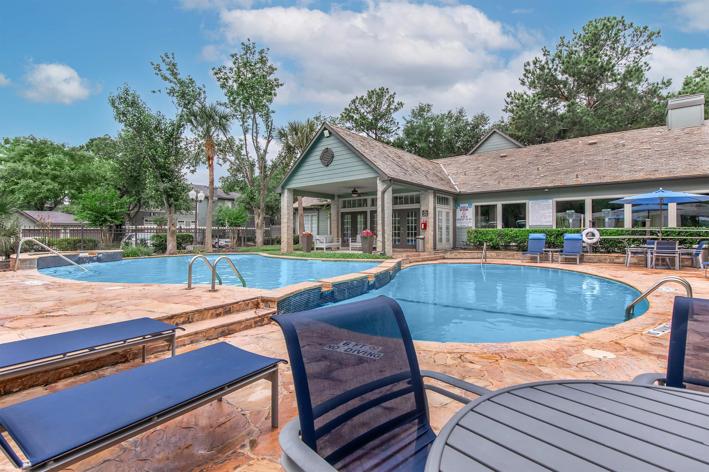 A clear swimming pool surrounded by a stone deck, featuring lounge chairs and an outdoor seating area. In the background, there is a building with large windows and greenery, including trees and plants, creating a serene ambiance. The sky is partly cloudy, enhancing the inviting atmosphere.