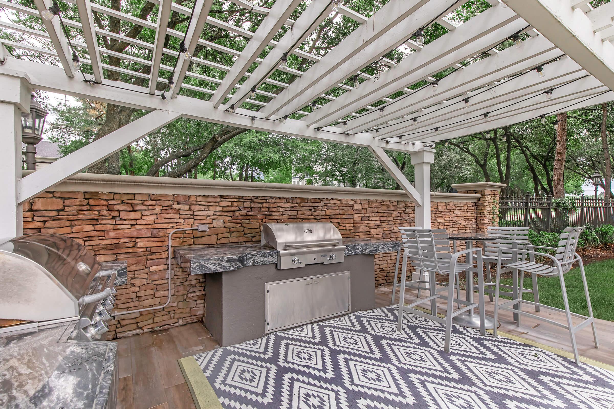 A backyard patio featuring a stylish outdoor kitchen with a stainless steel grill, a marble countertop, and modern bar stools. A decorative rug adds warmth to the space, surrounded by greenery and a stone wall, all under a white lattice pergola providing shade.