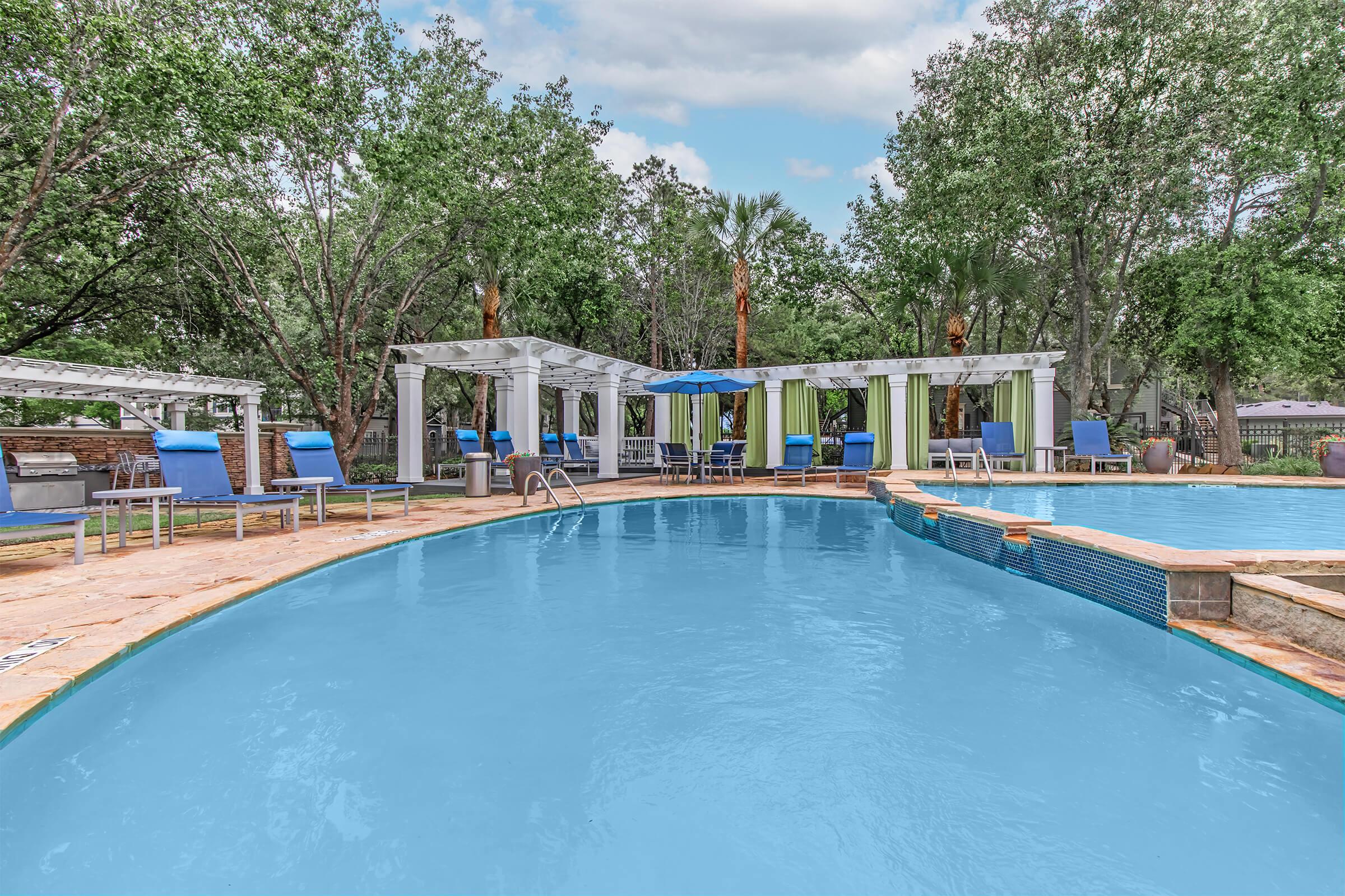 A tranquil pool area with bright blue water surrounded by lush greenery. There are several lounge chairs arranged around the pool, and shaded cabanas with curtains are visible in the background. The scene conveys a relaxing outdoor atmosphere ideal for leisure and recreation.