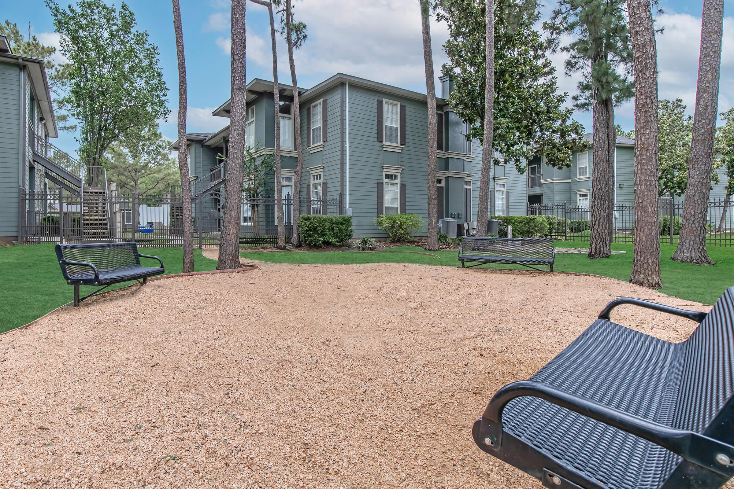 A gravel courtyard surrounded by trees, with two black benches positioned on either side. In the background, a multi-story building features gray siding and large windows. Lush green grass and bushes line the area, creating a peaceful outdoor space.