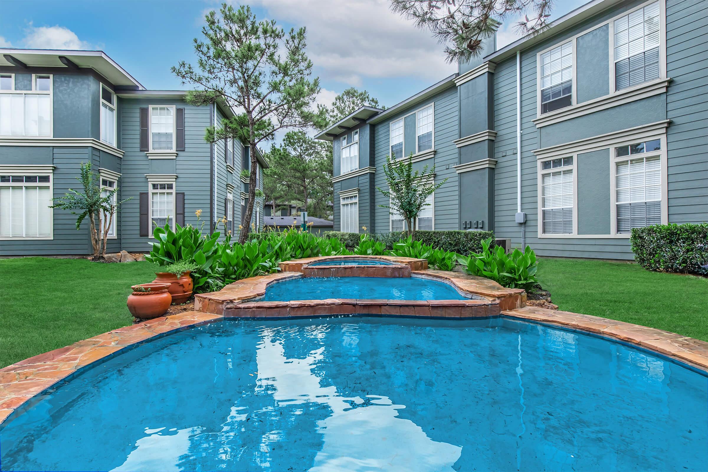 A serene outdoor scene featuring a clear blue swimming pool with a waterfall feature, surrounded by lush green plants and landscaping. The view includes two multi-story gray apartment buildings in the background, showcasing a calm and inviting atmosphere.