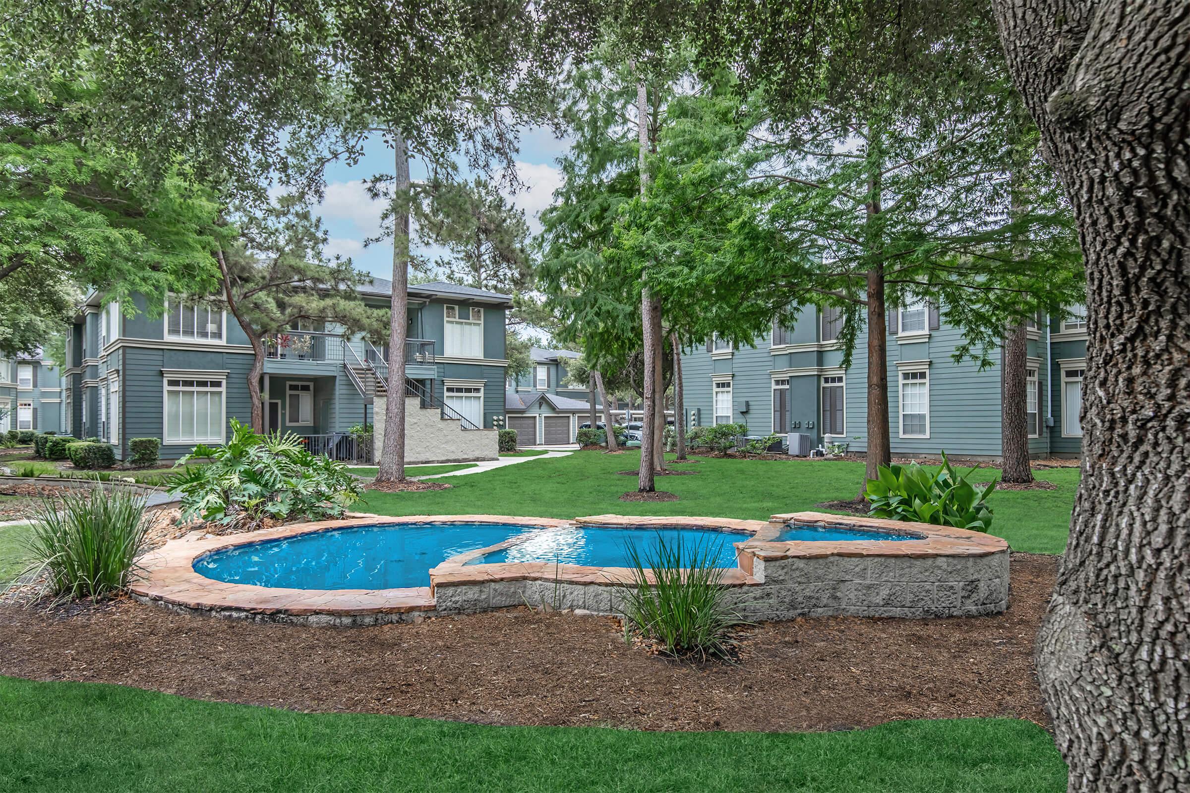 A well-maintained outdoor area featuring a bright blue swimming pool surrounded by landscaped greenery, including trees and bushes. Nearby, there are multi-story residential buildings with large windows, creating a serene atmosphere in a residential community.