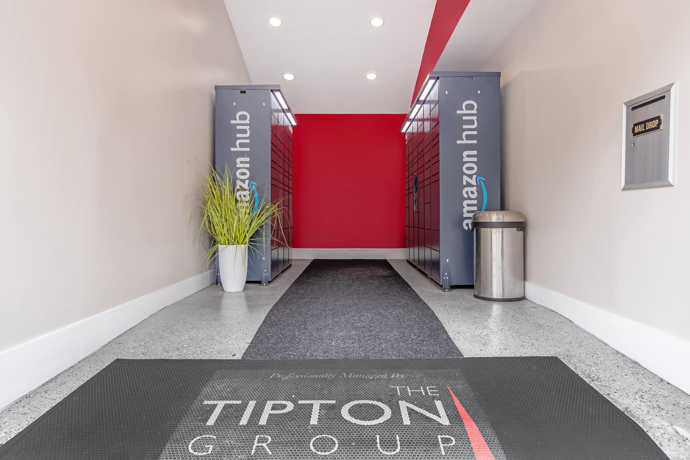 Interior entrance of a building featuring two Amazon Hub lockers on either side, a decorative plant in a white pot, and a trash bin. The floor has a black mat with "The Tipton Group" logo at the entrance, leading to a bright red wall with ceiling lights illuminating the space.