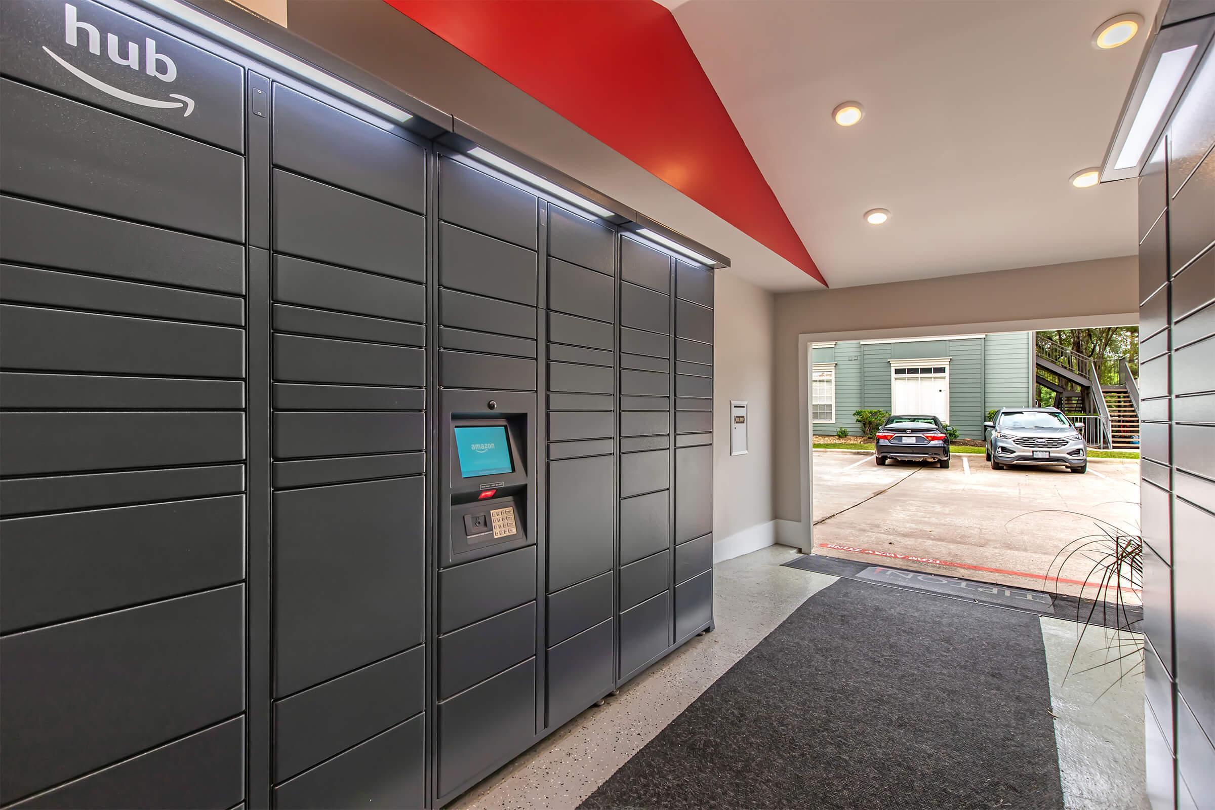 A modern entryway featuring secure delivery lockers labeled "hub." The lockers are gray and organized with a digital touchscreen interface for package retrieval. Visible in the background are parked cars and a well-lit, inviting interior leading outside.