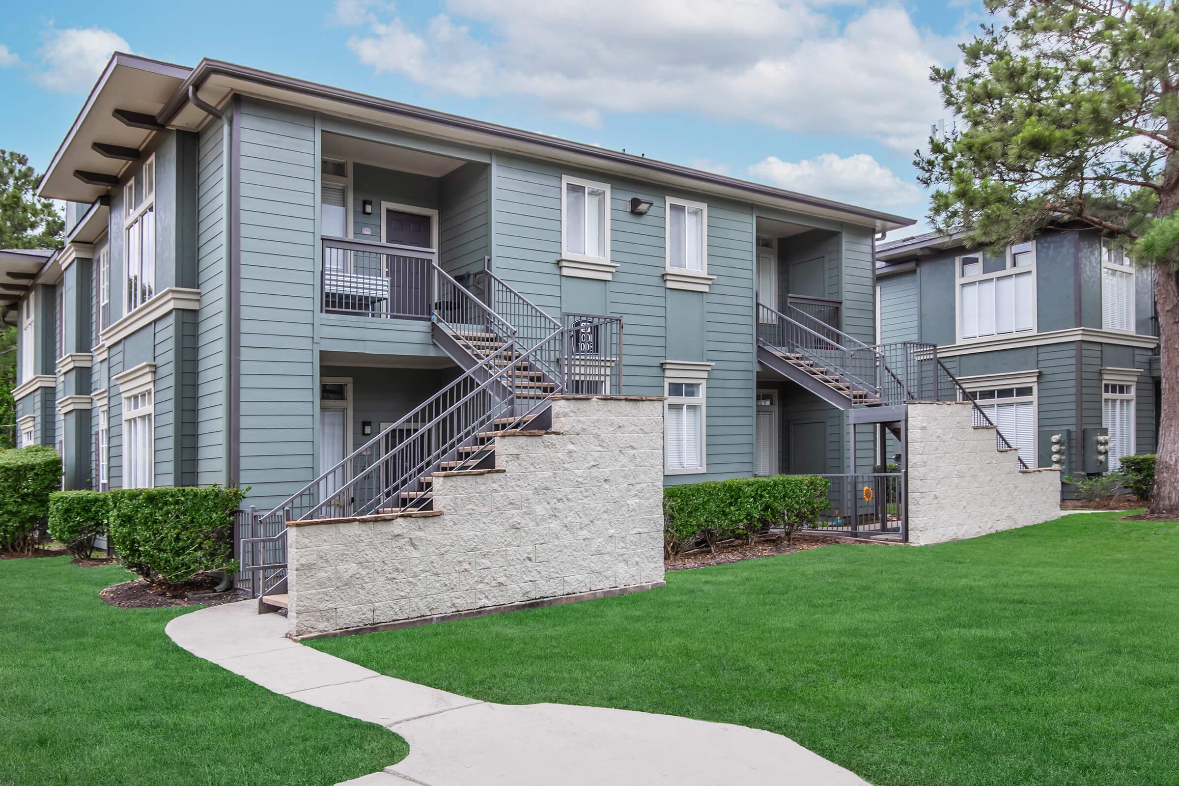 A two-story apartment building with a gray exterior, featuring stairs leading to the upper level. The building is surrounded by well-maintained green grass and small shrubs. A walkway leads up to the entrance, and blue skies with some clouds are visible in the background.