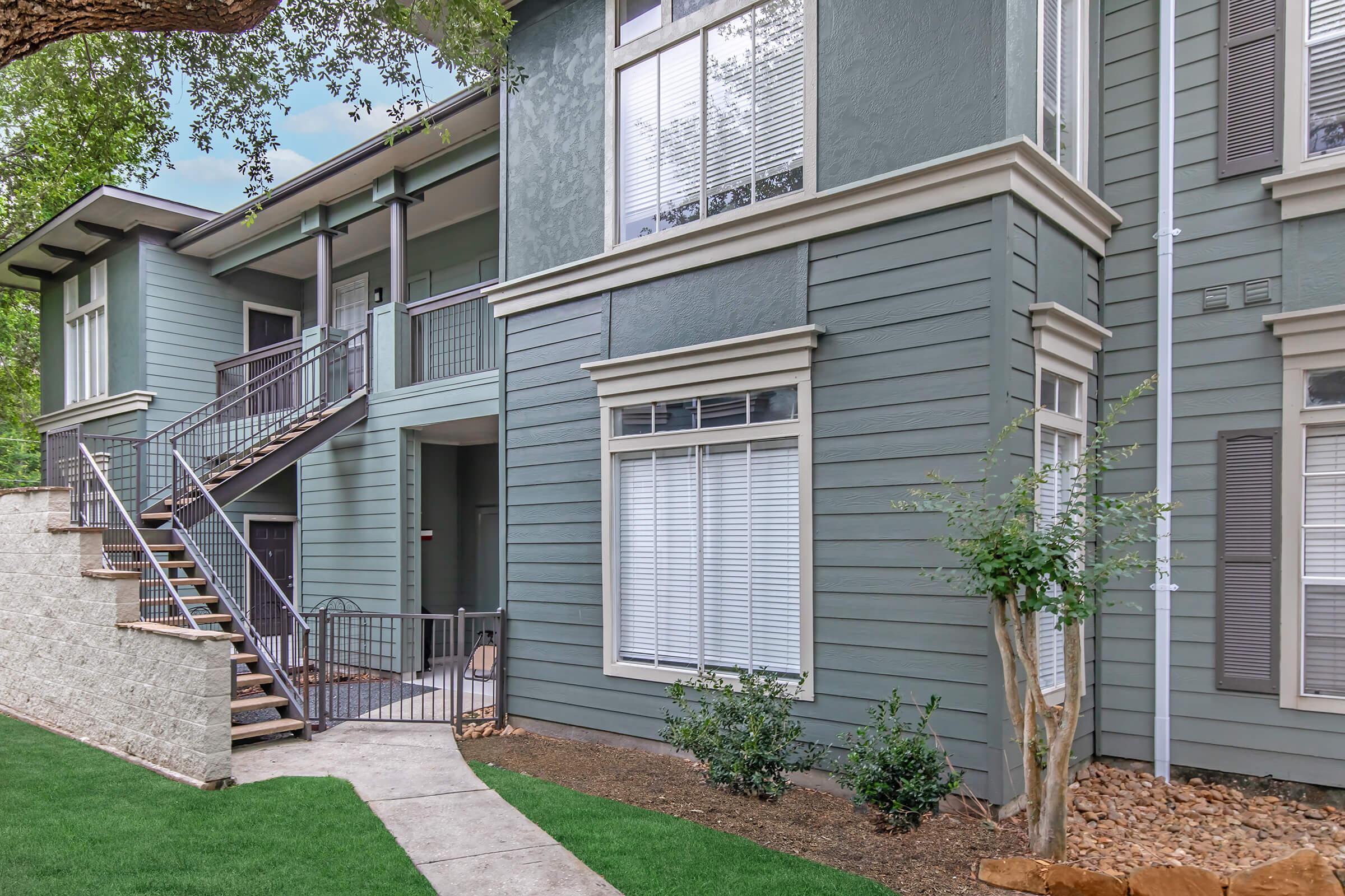 A modern apartment complex with green siding, featuring large windows and a stairway leading to upper units. There is a well-maintained lawn and landscaping, with a sidewalk leading to the entrance. The building's exterior has a contemporary design, complemented by surrounding trees.