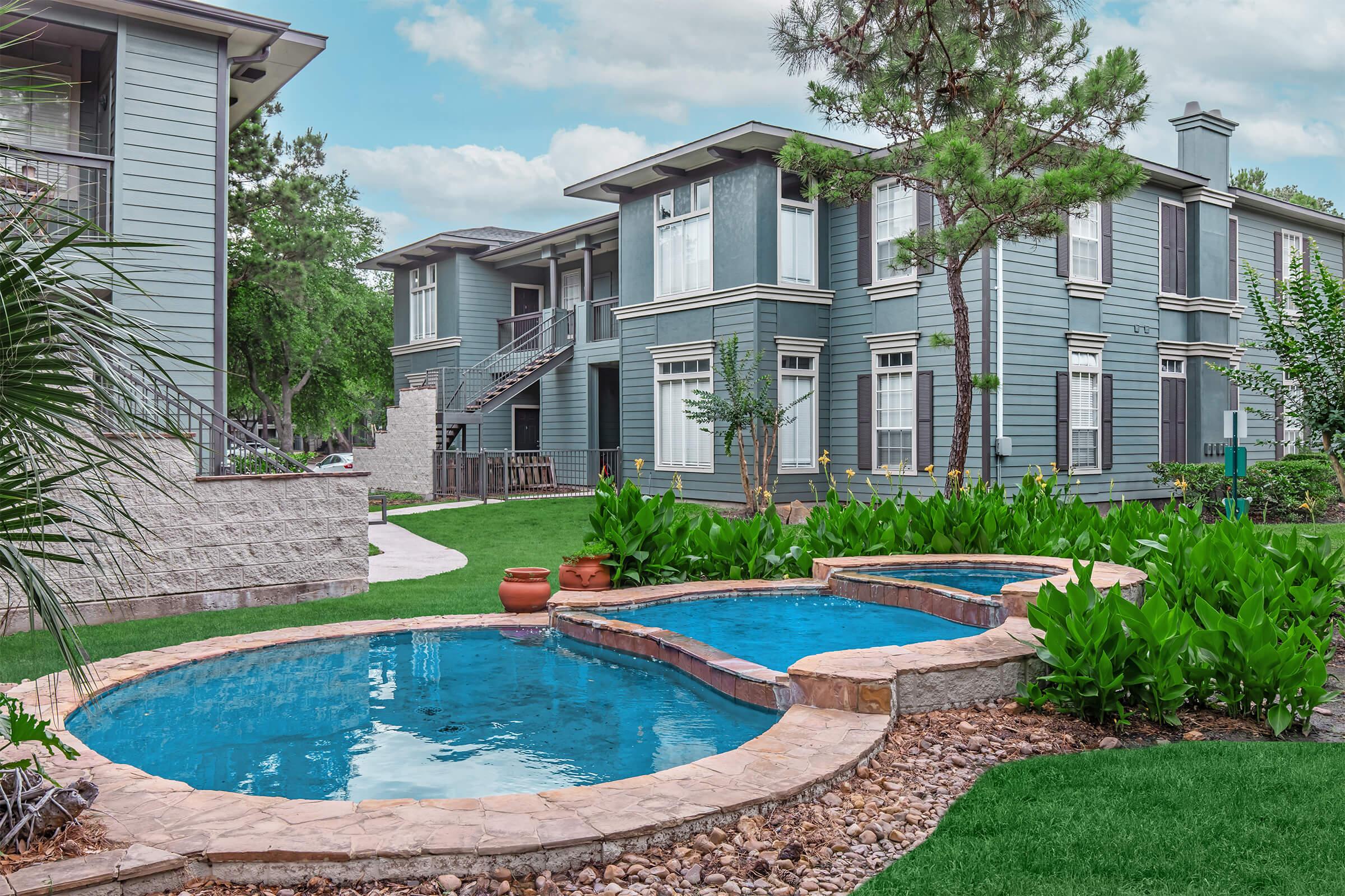 A peaceful courtyard featuring a blue, winding pool surrounded by lush green plants. The background shows modern two-story apartment buildings with large windows and balconies, set amidst a well-maintained grassy area and trees, under a cloudy sky.