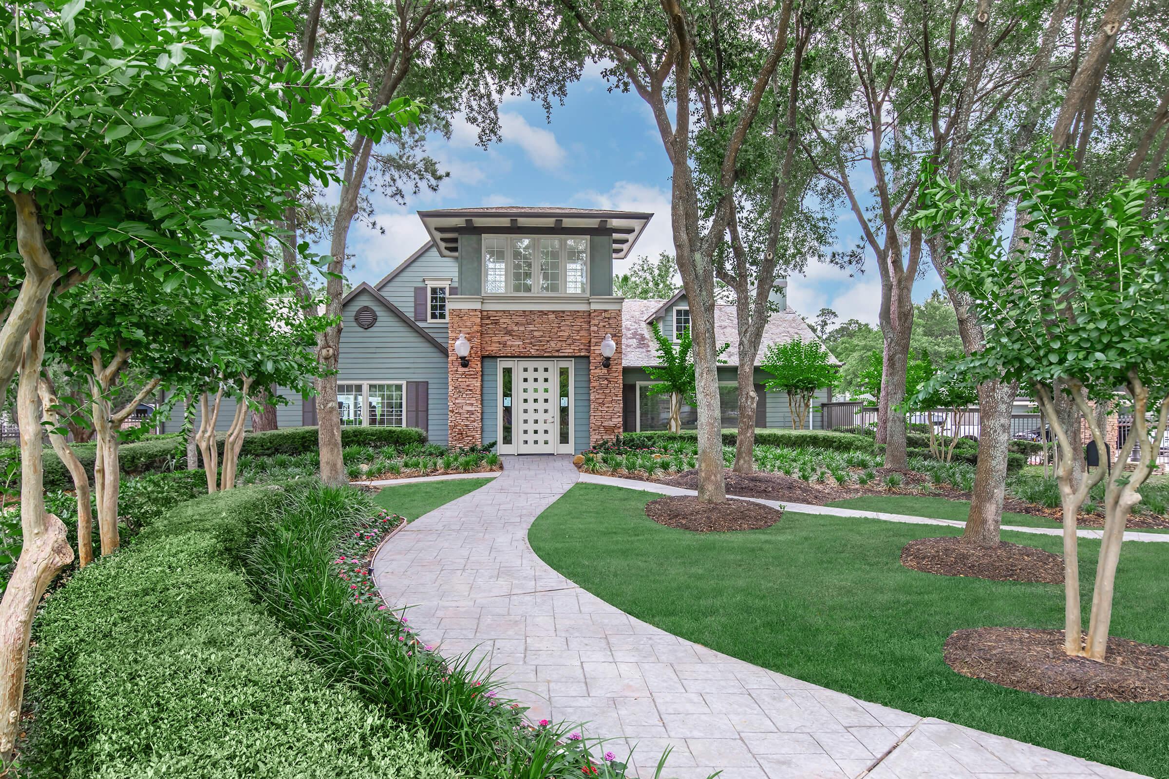 A modern two-story home with a mix of brick and siding, surrounded by lush greenery and neatly manicured lawns. The pathway leading to the front door is made of stone, flanked by flower beds and trees, creating an inviting entrance to the house.