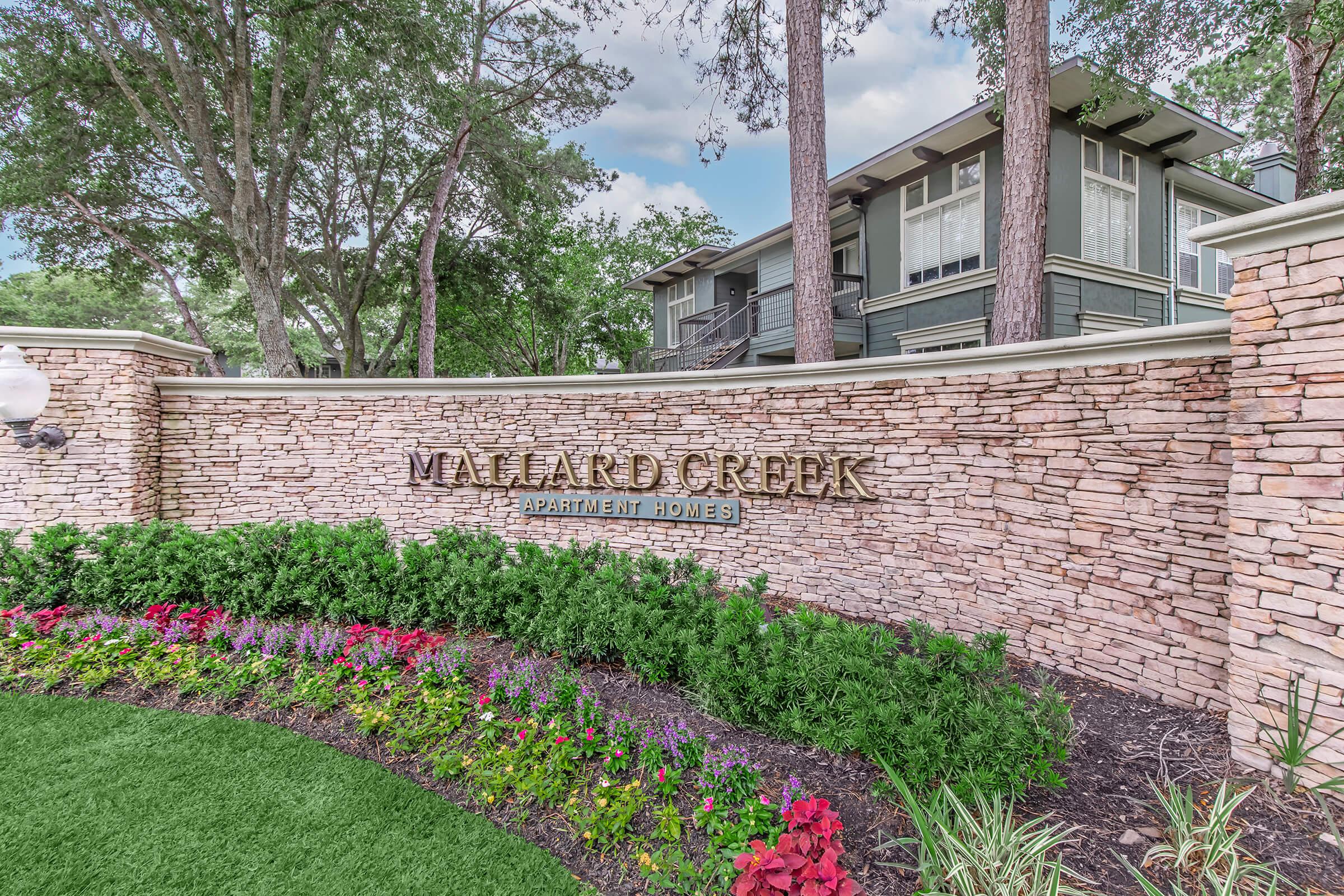 Stone entryway sign for Vallard Creek Apartment Homes, surrounded by colorful flowerbeds and lush greenery. The sign features the name "Vallard Creek" prominently, set against a backdrop of trees and an apartment building in the background.