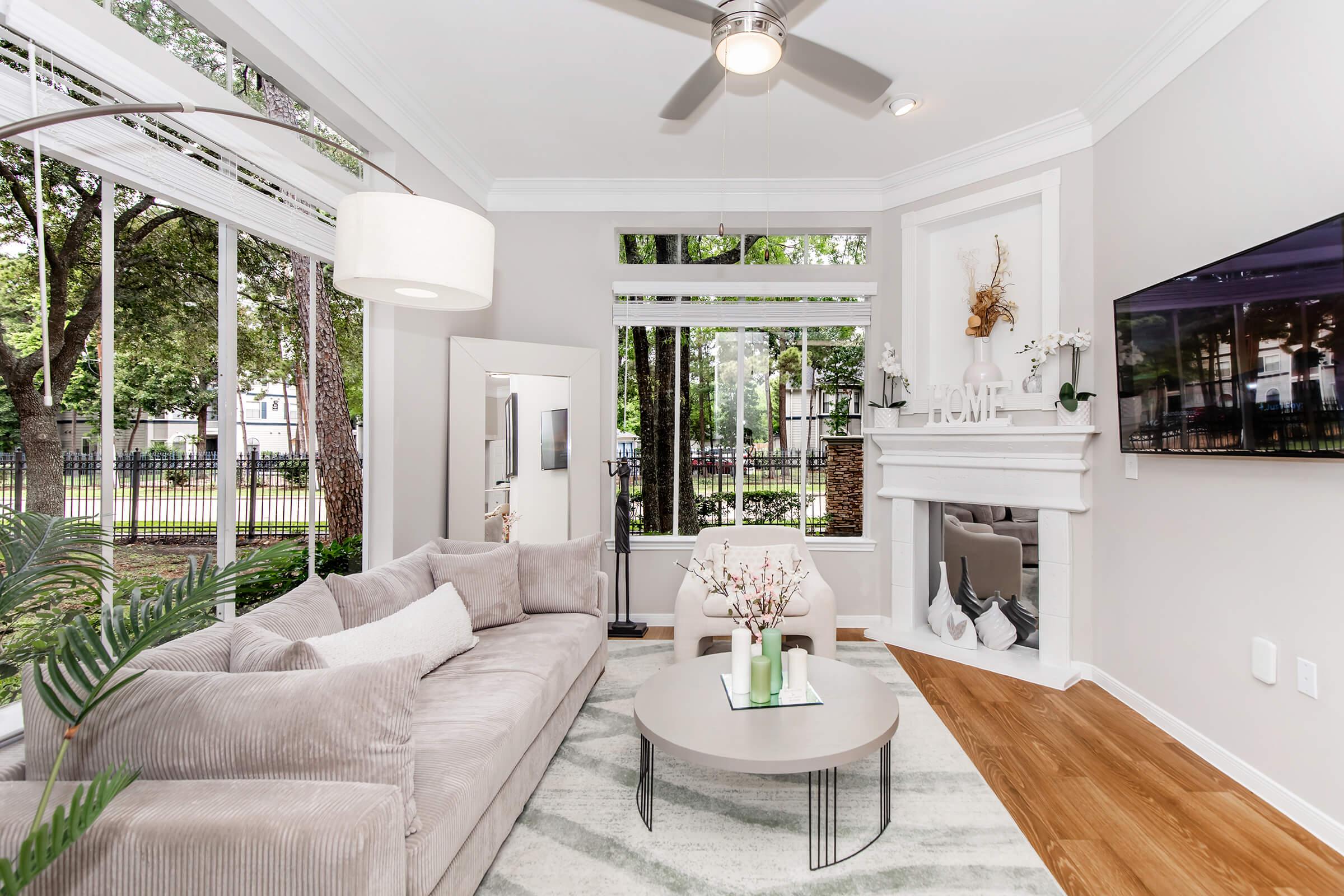 A cozy living room featuring a light gray sofa with decorative pillows, a round coffee table, and a stylish ceiling fan. Large windows allow natural light to illuminate the space, showcasing greenery outside. A modern TV is mounted on the wall above a decorative mantel, creating a welcoming atmosphere.