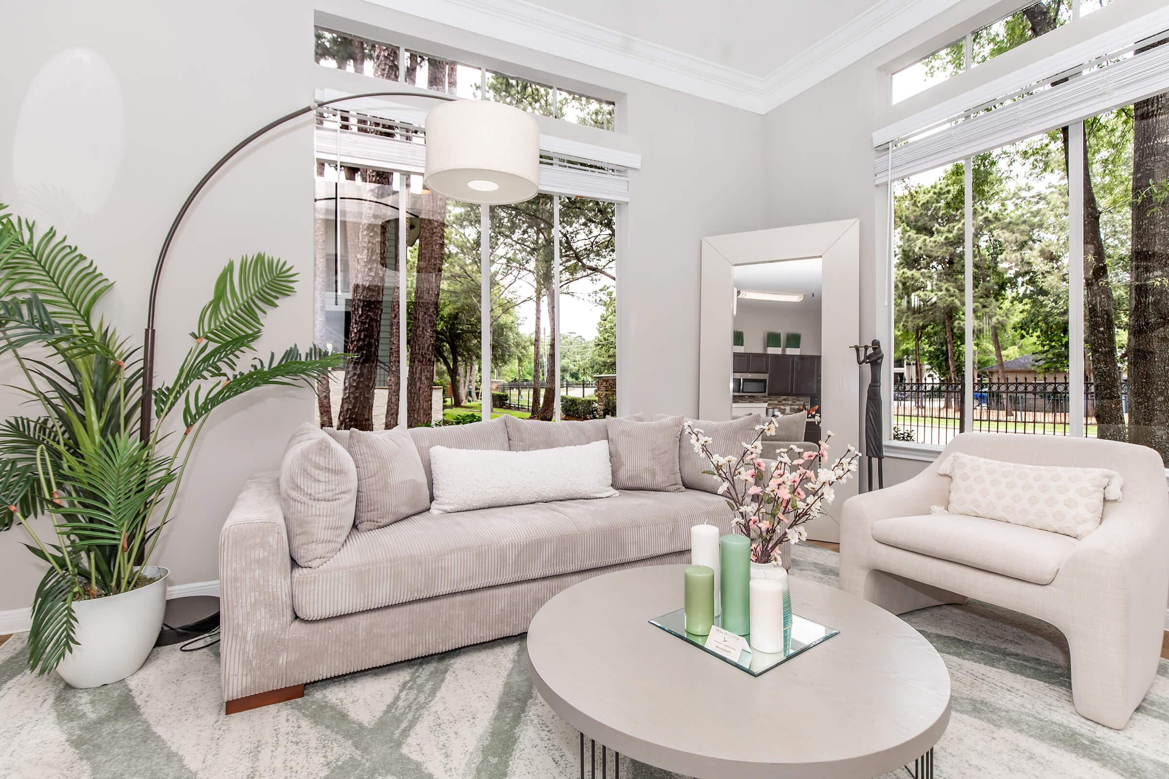 A modern living room featuring a light gray sectional sofa with decorative cushions, a round coffee table with candles and flowers, and a chic armchair. Large windows allow natural light, framing a view of greenery outside. The walls are painted a soft neutral color, enhancing the airy atmosphere.