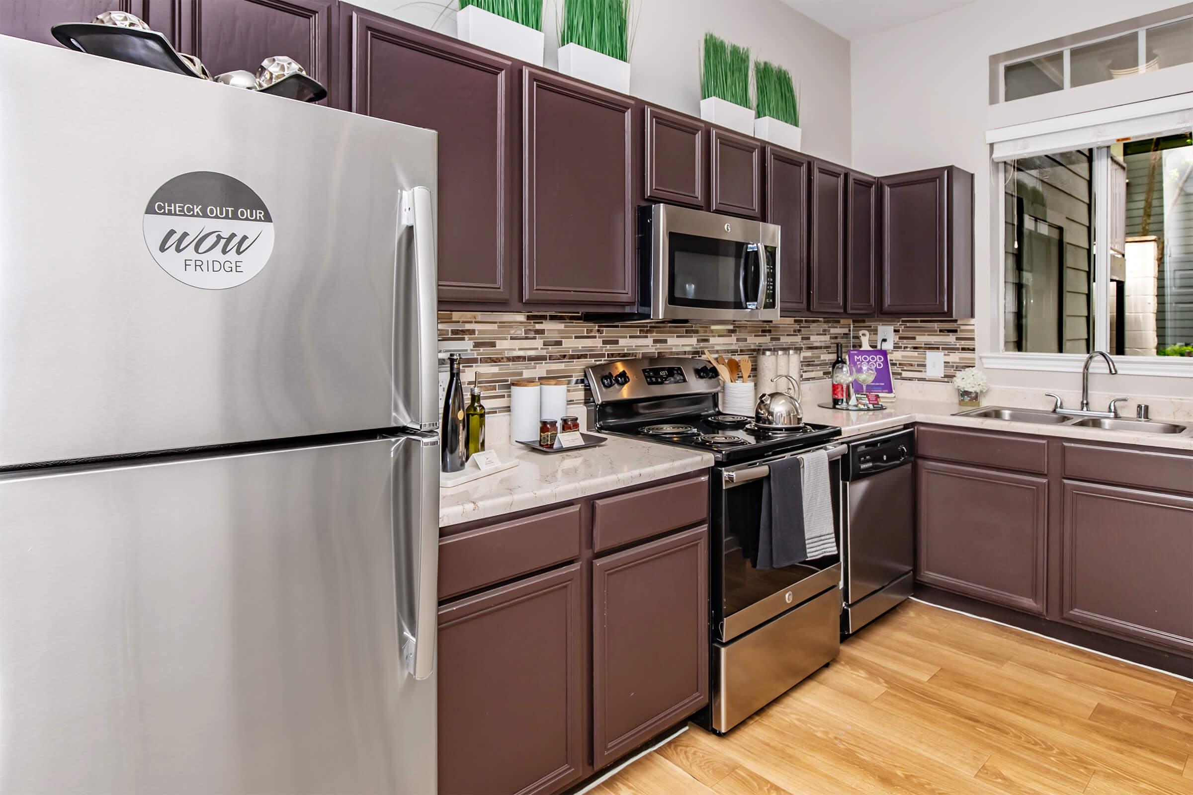 A modern kitchen featuring dark wood cabinetry, stainless steel appliances, and a light countertop. The refrigerator has a circular sticker saying "Check Out Our Wow Fridge." There are decorative plants on shelves, and the kitchen has a window providing natural light. The flooring is wooden.