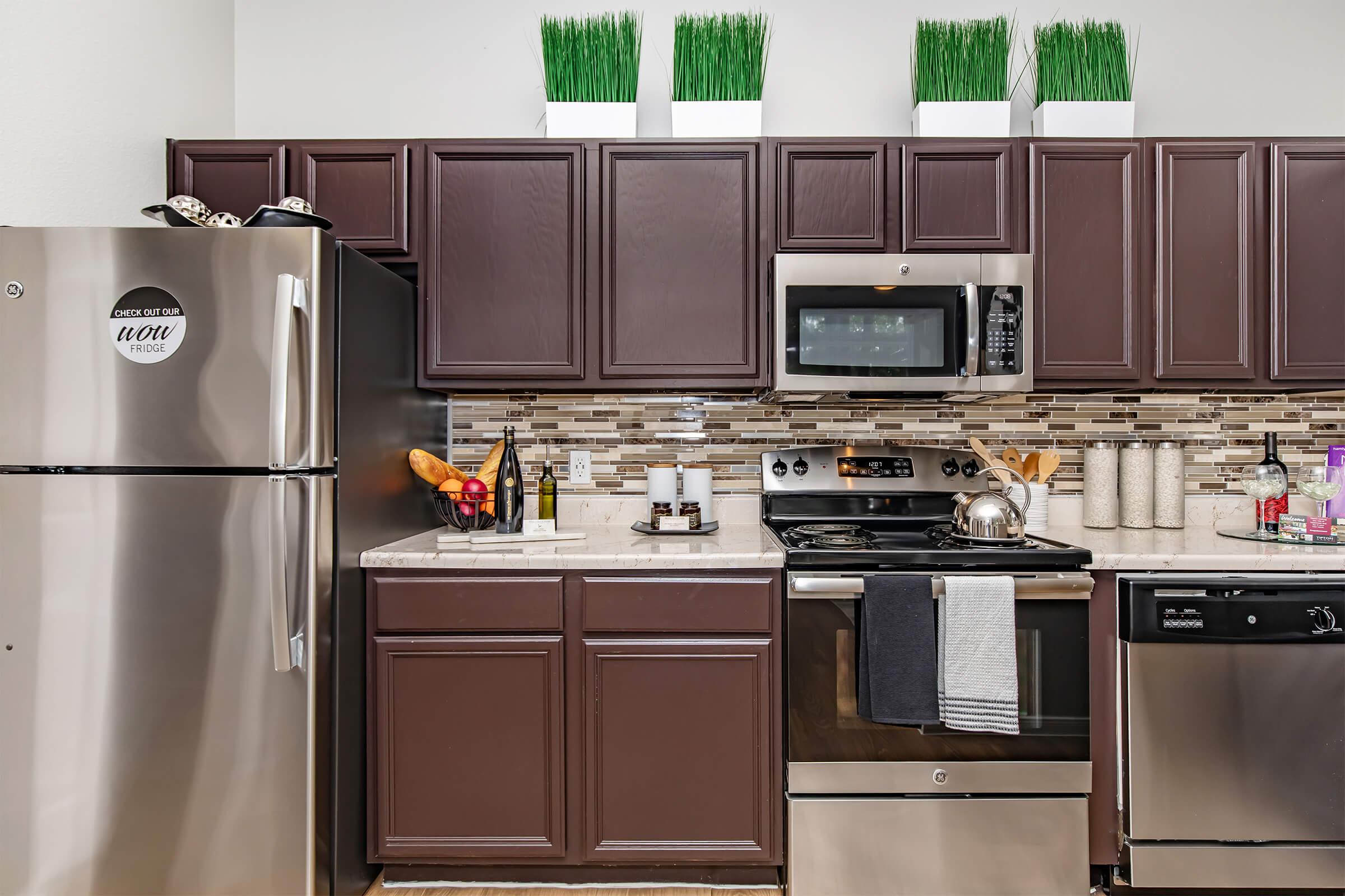 Modern kitchen featuring dark brown cabinets, stainless steel appliances including a fridge, oven, and dishwasher. A decorative backsplash and countertop items such as wine bottles and fruit add a stylish touch. Green planters with grass-like decor sit atop the cabinets, enhancing the overall aesthetic.