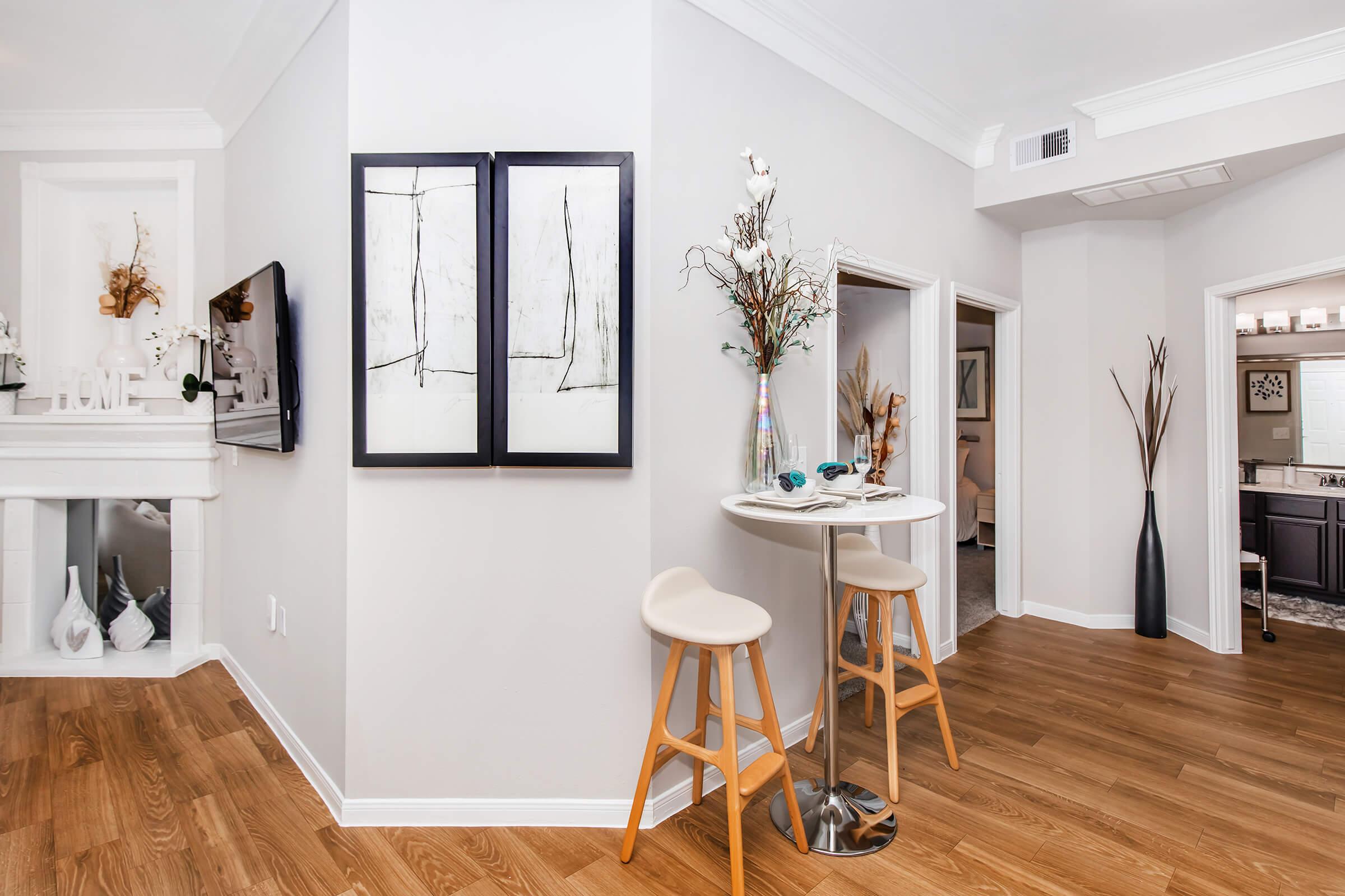 A modern interior featuring a light-colored wall with two framed artworks, a small round table with two stylish chairs, and a decorative vase on the table. In the background, there’s a fireplace and doorways leading to other rooms. The flooring is wooden, contributing to a warm, inviting atmosphere.