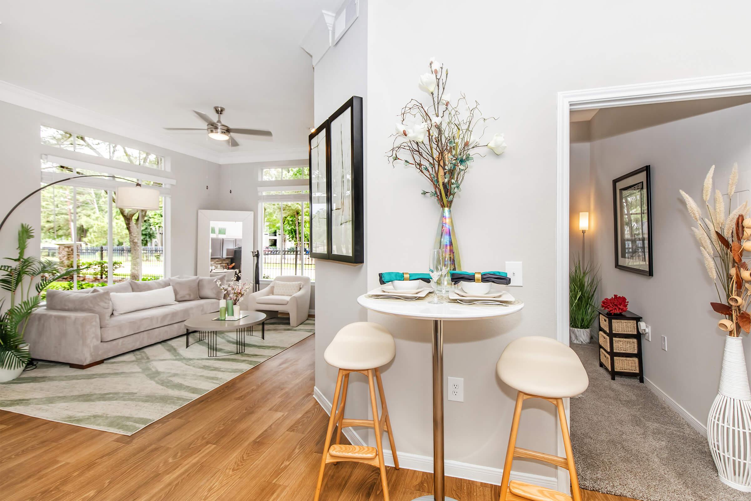 A modern living space featuring a cozy seating area with a light gray sofa and a round coffee table. Nearby, a small dining table with two stools sits beside a decorative vase. Large windows allow natural light, and a doorway leads to a carpeted room with decorative plants. The decor is stylish and inviting.