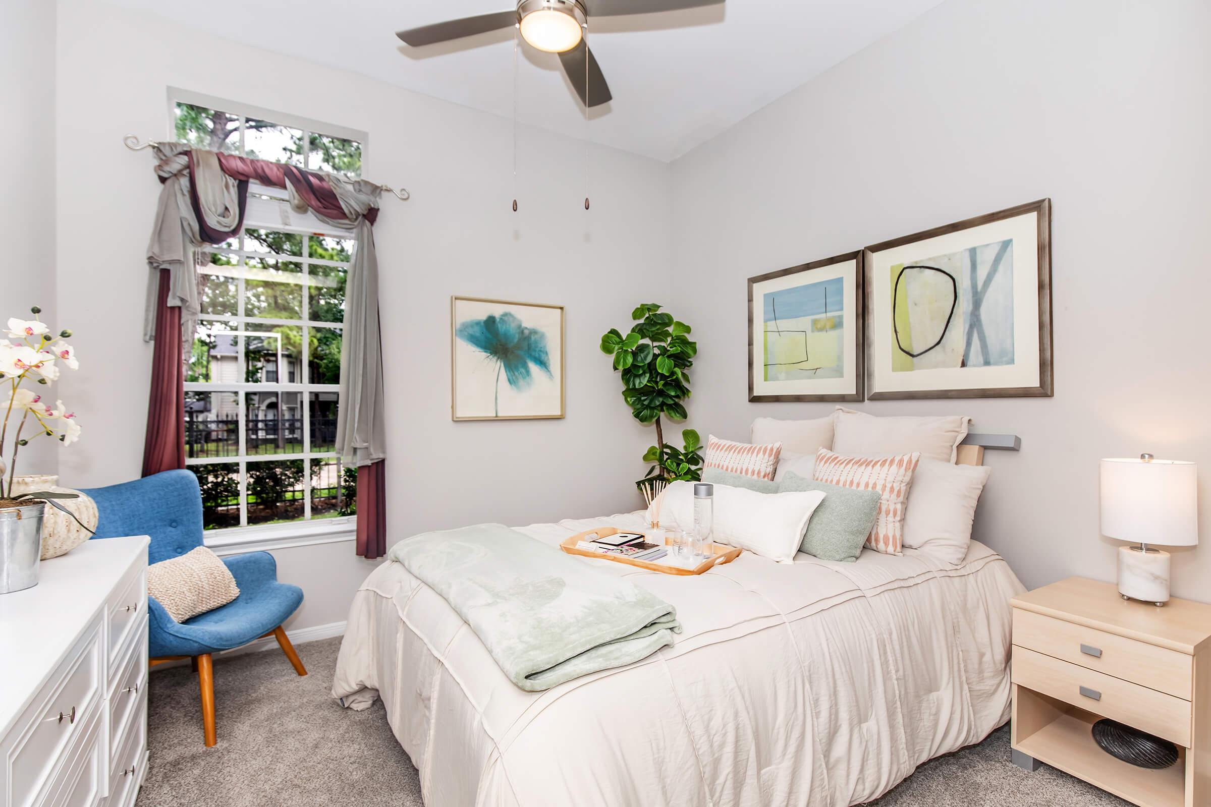 A cozy bedroom featuring a bed with light-colored bedding, decorative pillows, and a bedside table with a lamp. A blue upholstered chair is placed nearby, alongside a window with natural light. Wall art and a potted plant add a touch of style to the space, complemented by soft gray carpet and neutral wall colors.