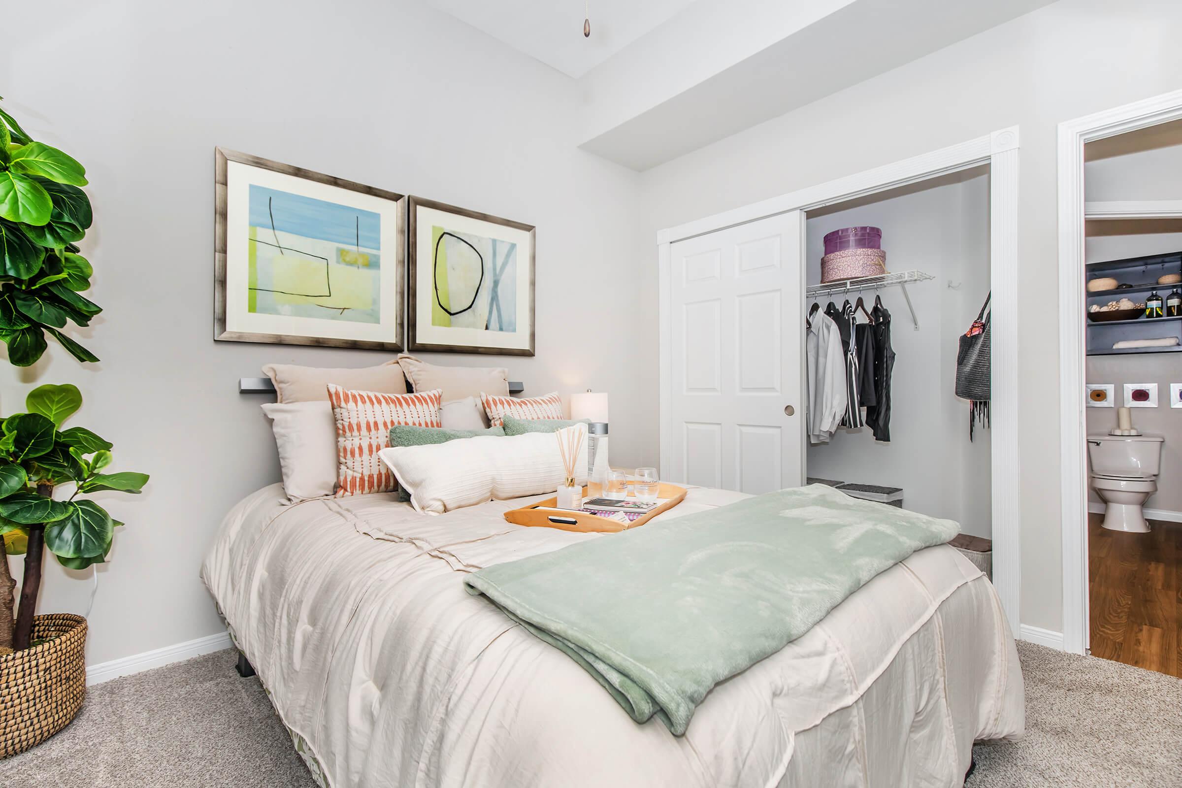 A cozy bedroom featuring a neatly made bed with decorative pillows and a light green throw. There are two framed artworks above the bed, a bedside table with a lamp, and a plant in the corner. An open closet displays clothing, and a bathroom door is visible in the background, indicating a well-organized space.