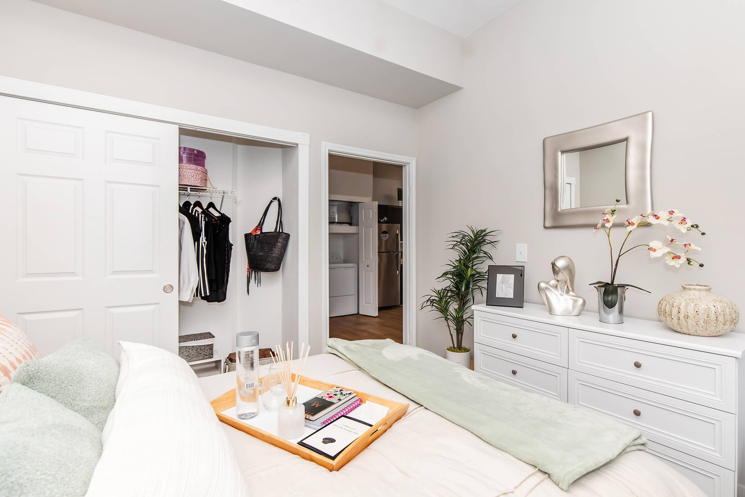 A cozy bedroom featuring a neatly made bed with soft pillows and a light green throw. A white dresser with decorative items and a plant is visible. An open closet shows clothes and a couple of bags, while a mirror hangs on the wall. The room has warm lighting and a soft color palette.