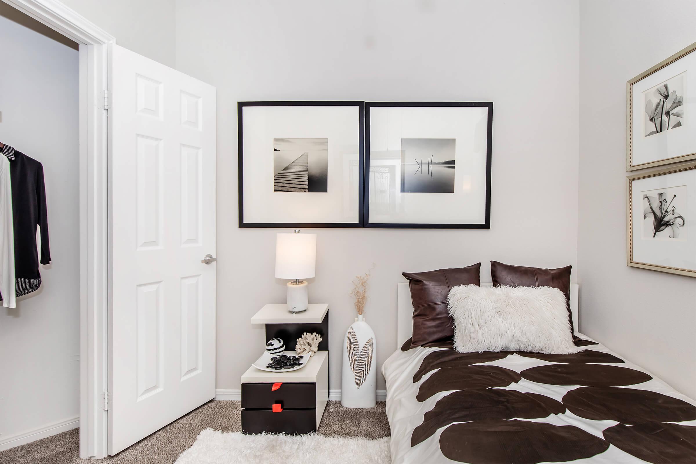 A cozy bedroom featuring a single bed with a polka dot comforter, a lamp on a nightstand, and decorative art on the walls. There’s a white vase and a small dark storage box beside the bed, and a closet door is visible in the background. Soft carpet and neutral wall colors create a warm ambiance.