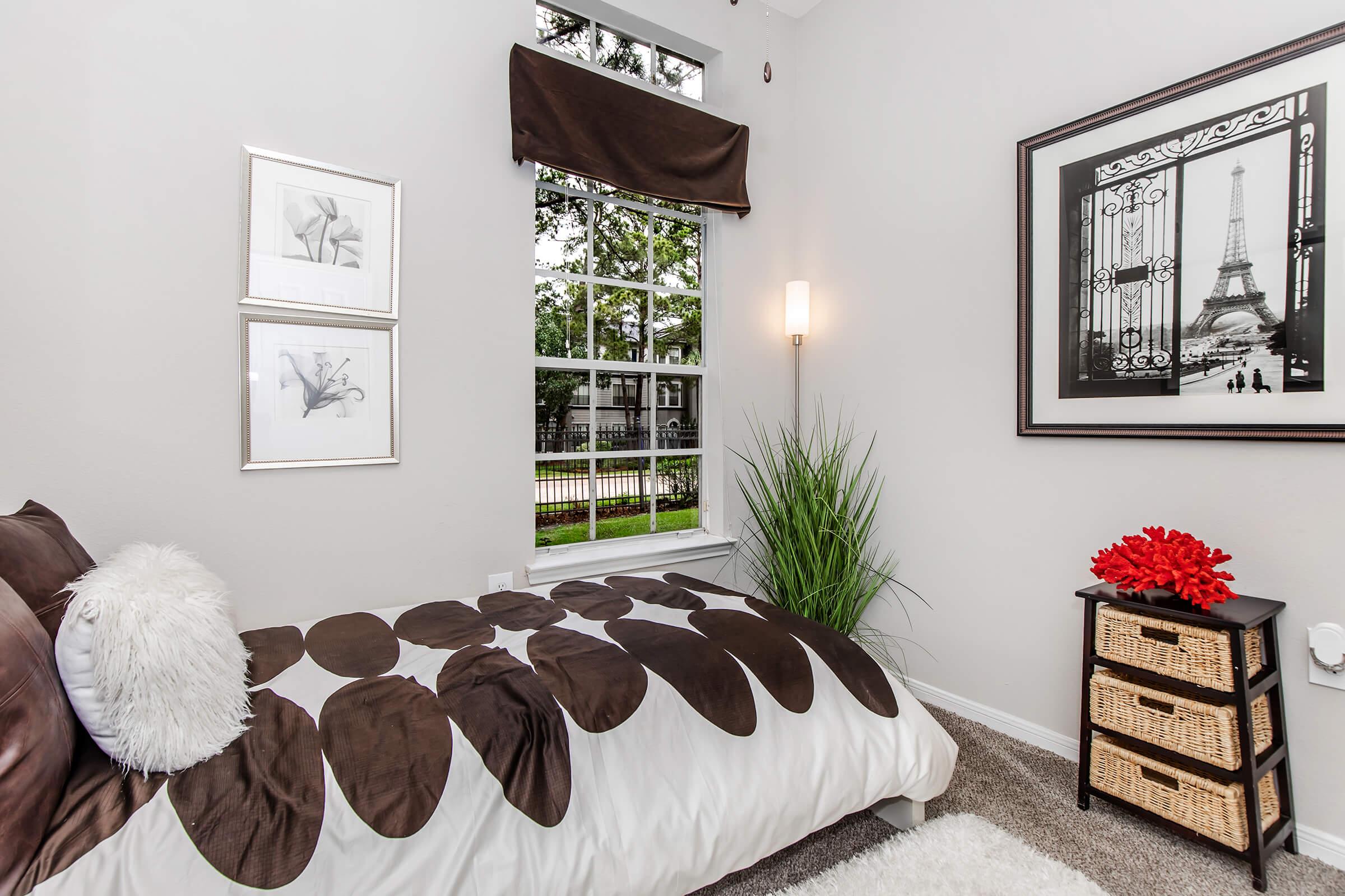 Cozy bedroom featuring a bed with a patterned duvet cover, a decorative pillow, and a bedside lamp. There's a large window with a view of greenery, framed black-and-white artwork of the Eiffel Tower on the wall, and two framed pictures. A small wicker storage basket sits next to the bed, adding to the room's charm.