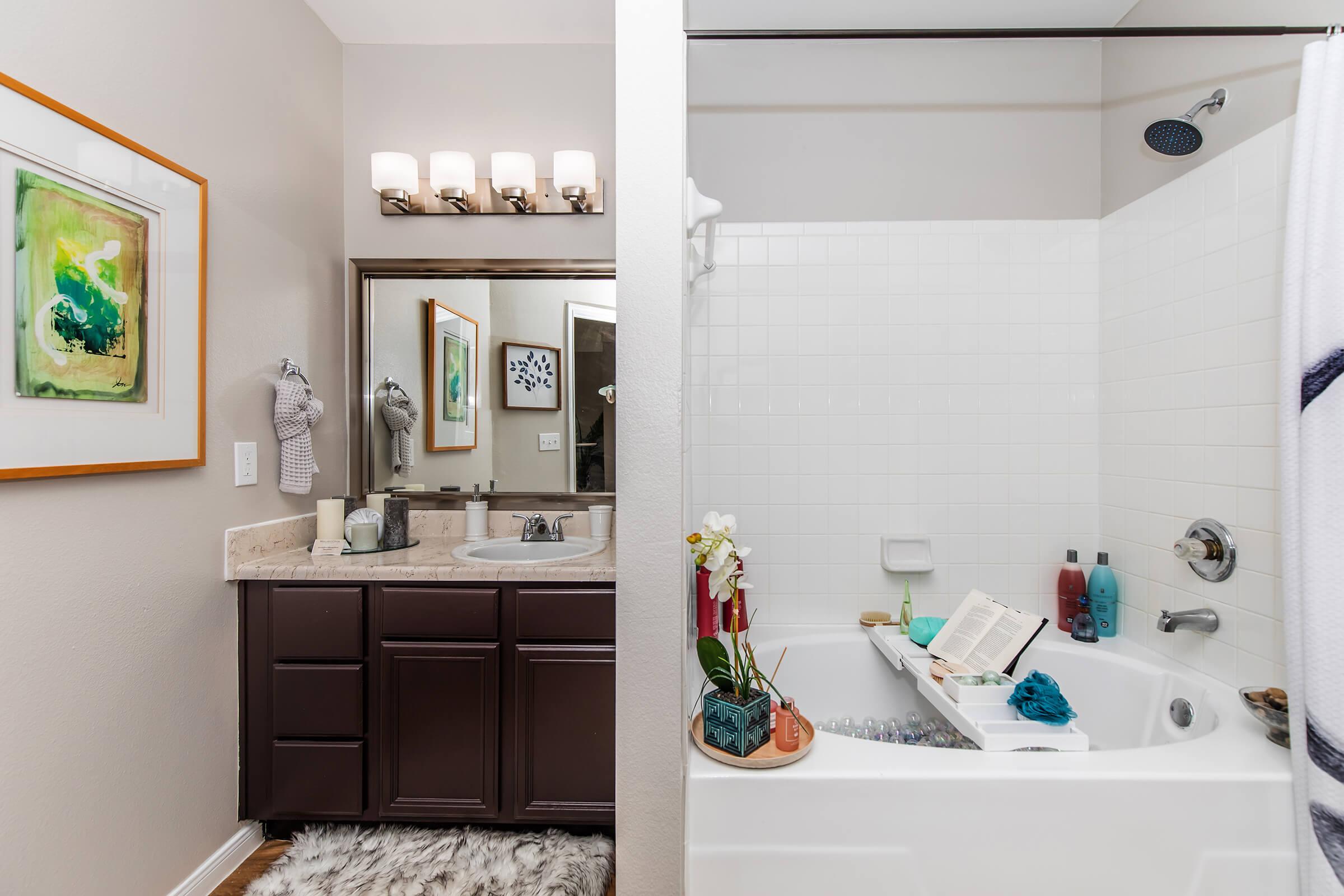 A well-lit bathroom featuring a double vanity with a large mirror, modern light fixtures, and dark cabinets. On the right, a white bathtub with a shower has a book resting on the edge, surrounded by decorative items and toiletries. Soft gray rugs add a touch of comfort to the space.