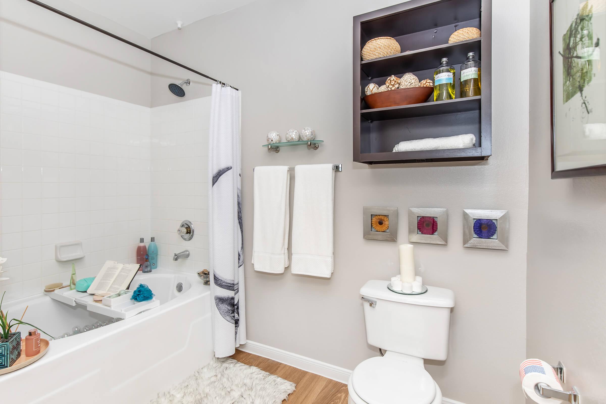 A modern bathroom featuring a bathtub with a shower, white towels hanging on a towel rack, and a shelf containing decorative items and toiletries. The walls are painted in a soft gray, and there is a small rug on the floor, adding a cozy touch to the space.