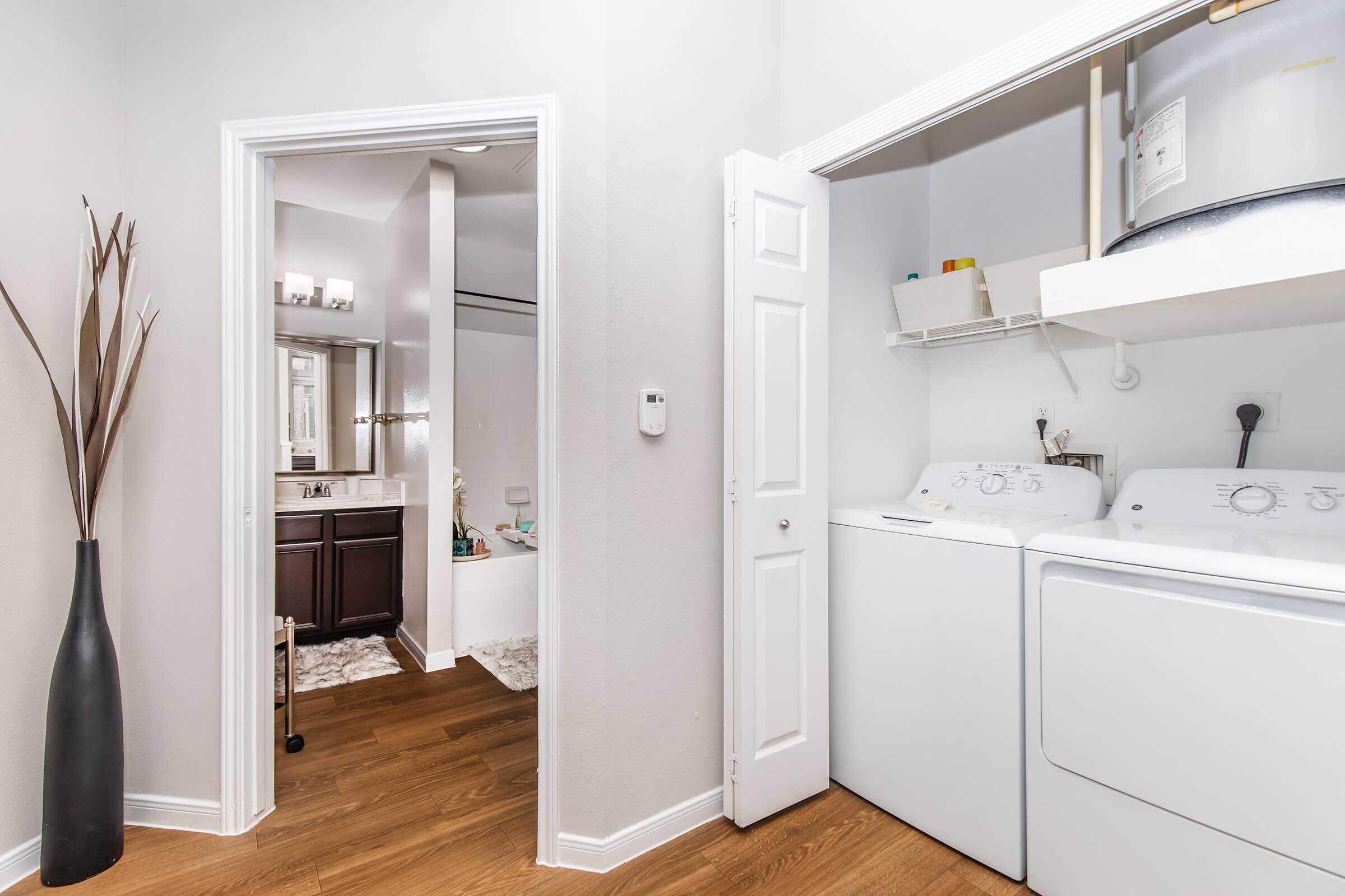 A modern laundry area with white washer and dryer, located next to a bathroom. The walls are light-colored, and there is a tall black vase with decor near the entrance. The flooring is wood, and a doorway leads to a well-lit bathroom with a tub and mirror visible.