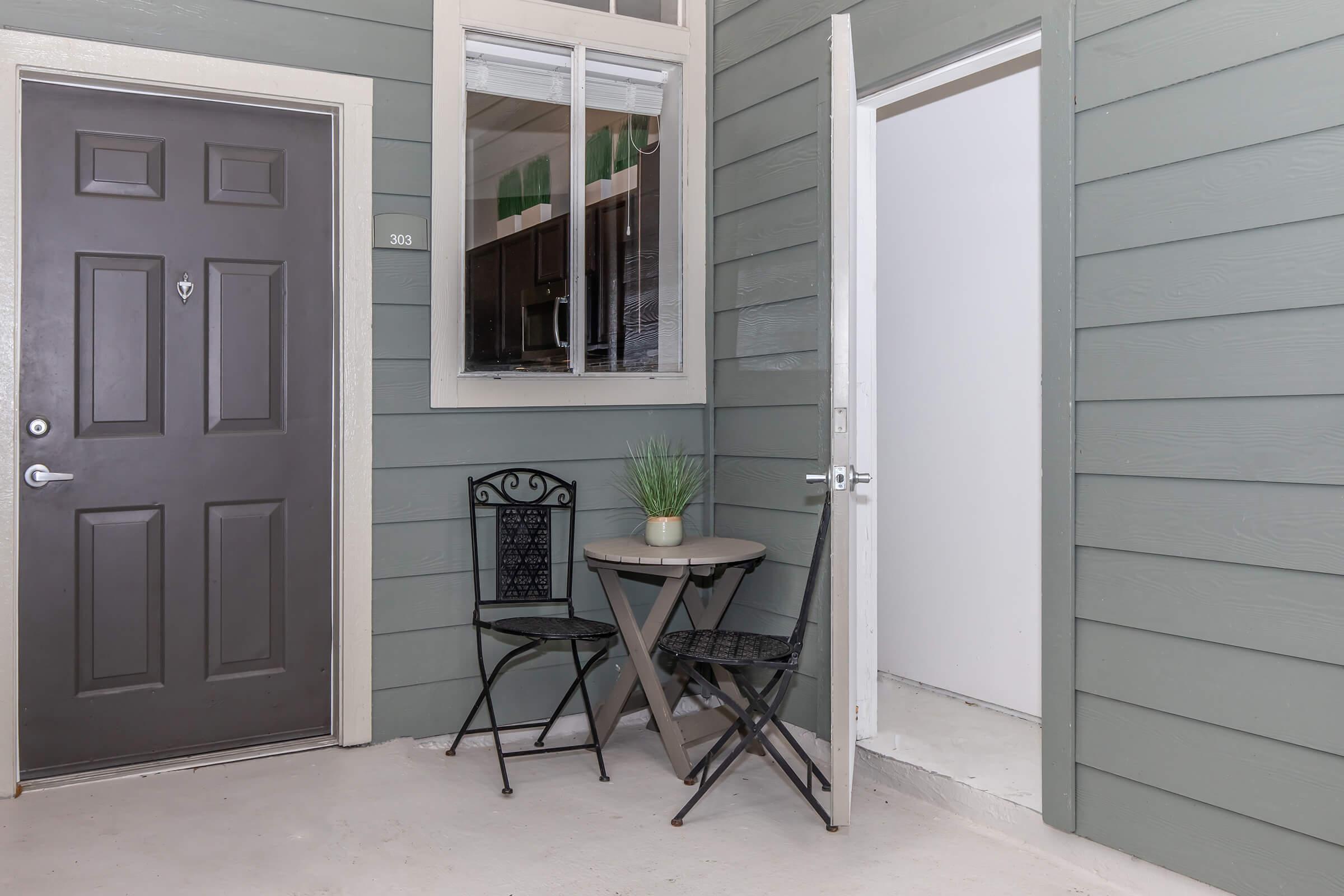 A small outdoor area featuring a dark door with the number 303, a round table with two black chairs, and a decorative plant on the table. Next to the door is an open entryway leading to a white interior space. The walls are painted in a muted green color, adding a modern touch to the entrance.