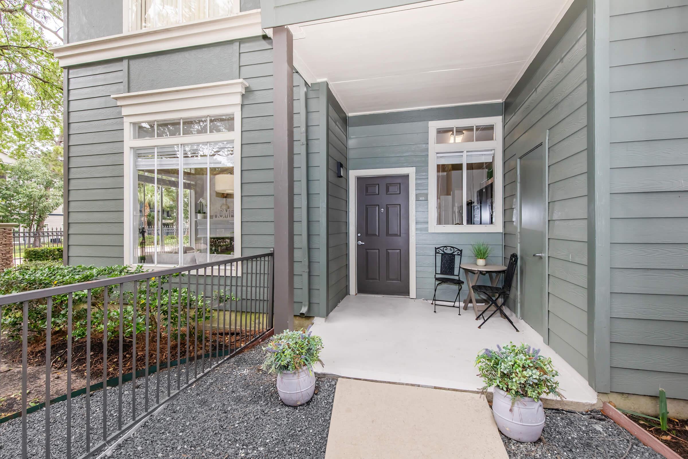 A modern entrance to an apartment featuring a small porch with a decorative rug, two potted plants, and a bistro table with chairs. The walls are painted in a soft green color, and large windows invite natural light, creating a welcoming atmosphere.