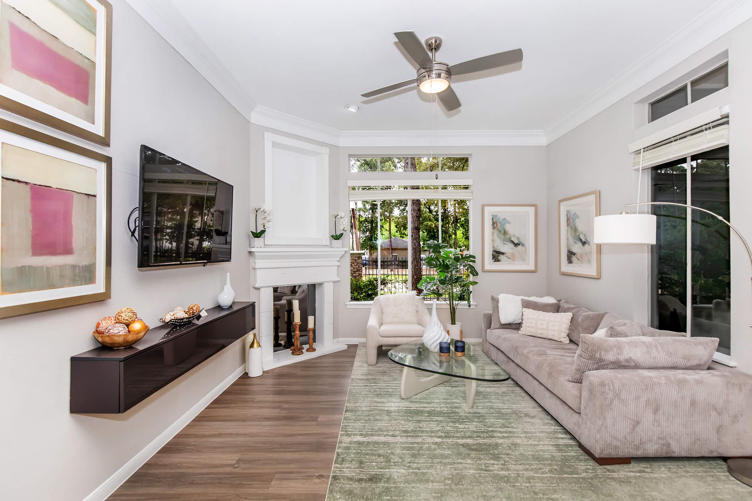 Cozy living room featuring a light gray sofa and glass coffee table on a green area rug. A modern ceiling fan is visible, along with a mounted television. Large windows let in natural light, and the decor includes abstract art and a stylish floor lamp. A fireplace adds warmth to the space, surrounded by greenery.