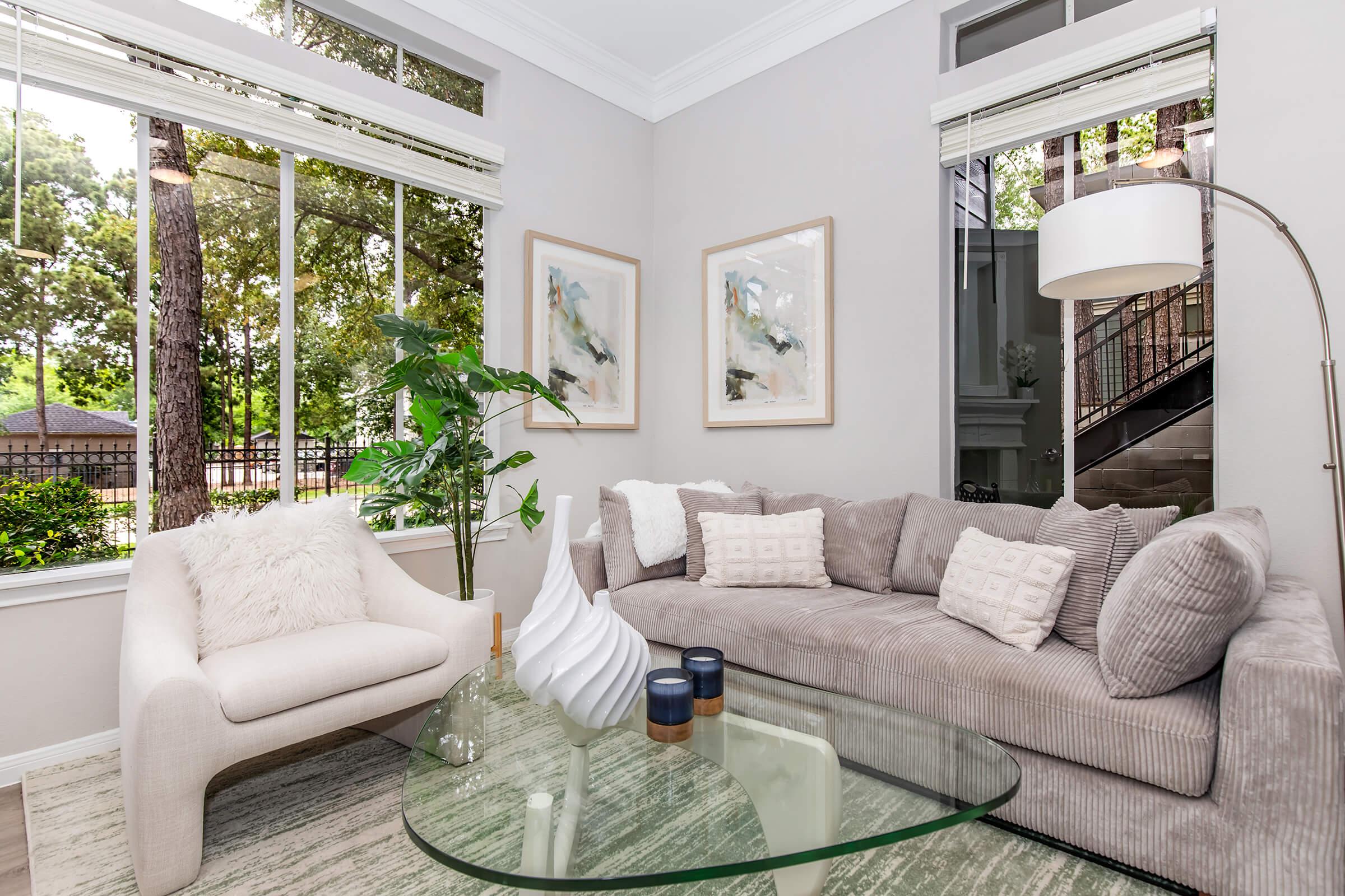 A cozy living room featuring a light gray sectional sofa with decorative pillows, a white armchair, and a glass coffee table. Natural light streams through large windows, illuminating the space and showcasing greenery outside. Two framed art pieces adorn the wall, adding a touch of elegance to the decor.