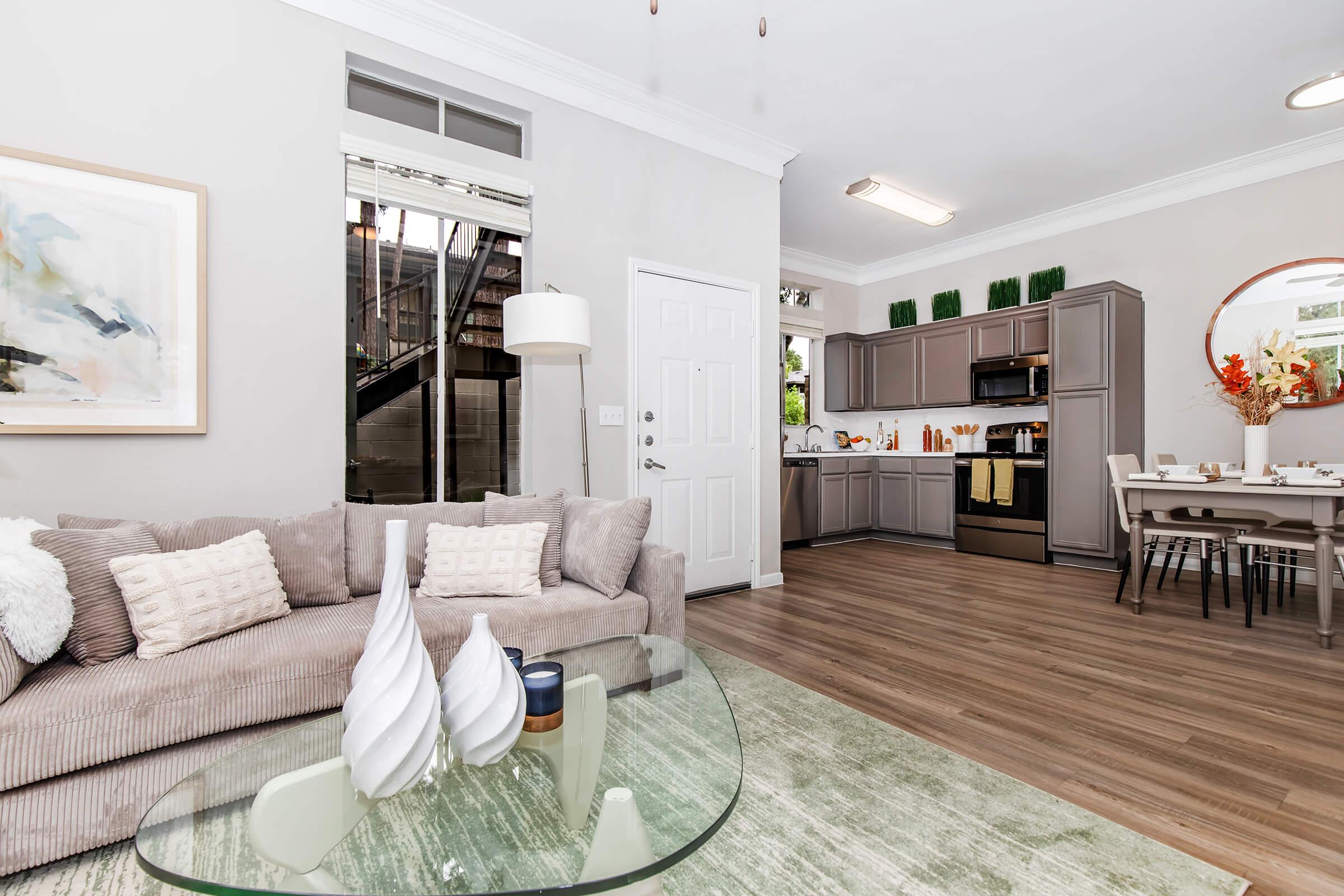 A modern living space featuring a cozy gray sofa with decorative pillows, a glass coffee table, and a dining area with a wood table and chairs. The kitchen in the background has gray cabinetry and stainless-steel appliances, with natural light coming through a window. Decorative plants and artwork enhance the ambiance.