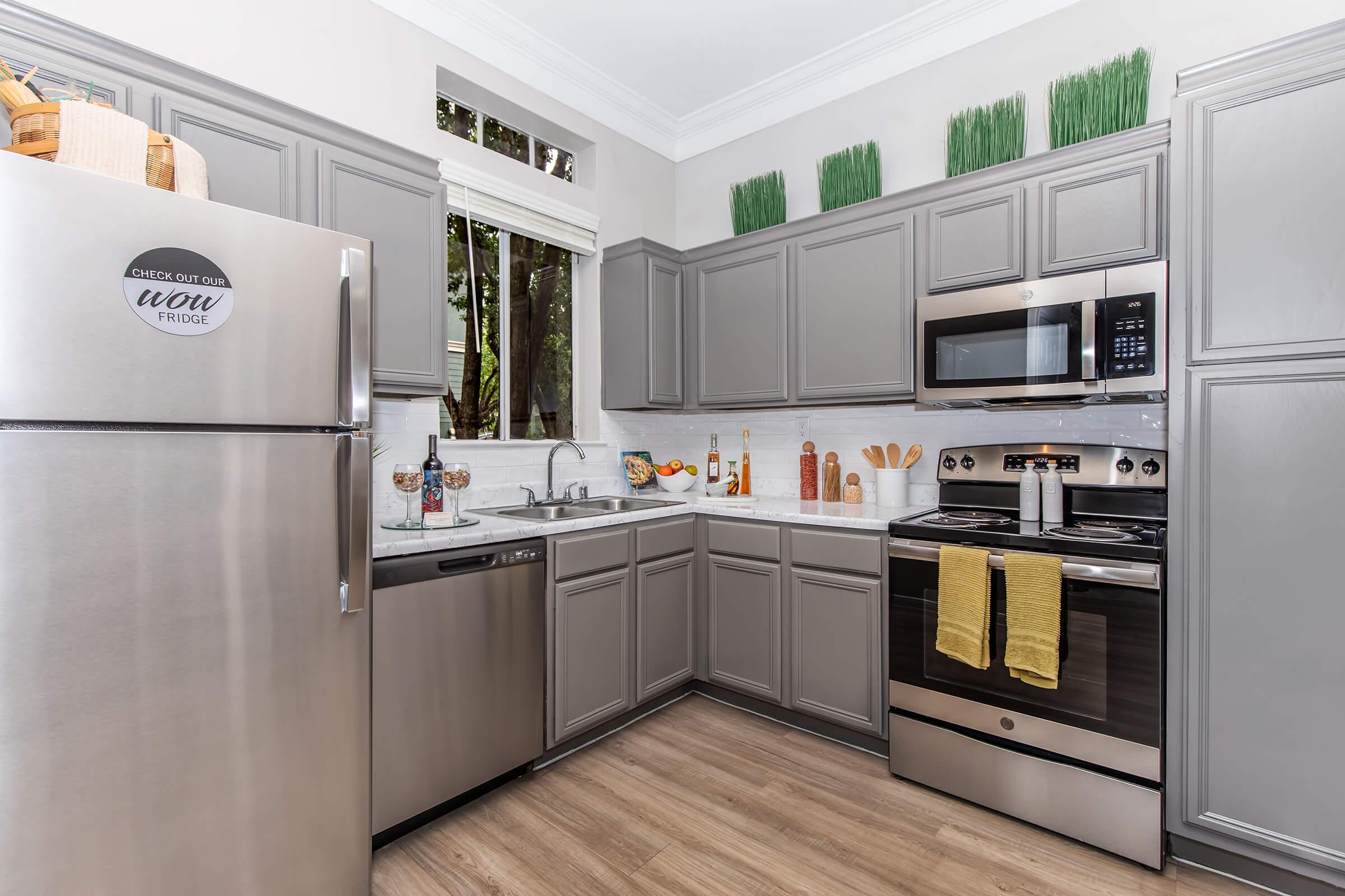 Modern kitchen featuring gray cabinets, stainless steel appliances including a refrigerator and oven, a double sink, and a dishwasher. The countertop is adorned with decorative bottles and cooking utensils. Natural light streams in from a window, enhancing the warm, inviting atmosphere.