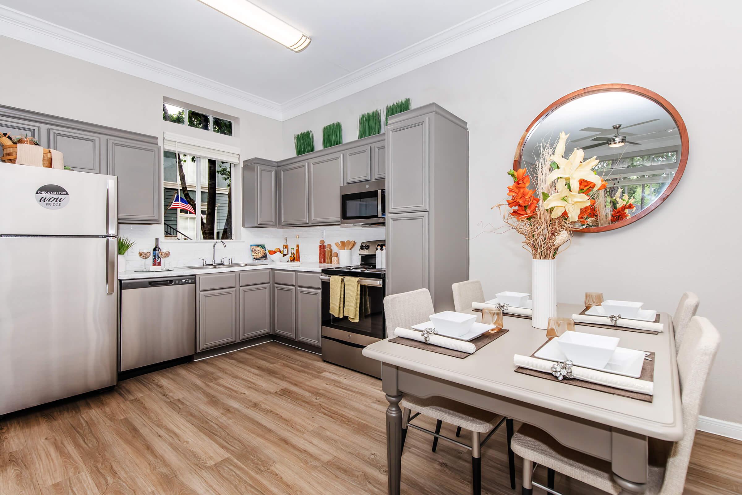 A modern kitchen with gray cabinetry and stainless steel appliances, featuring a dining area with a stylish table set for four. Decorative elements include a round mirror, a floral centerpiece, and greenery accents on the shelves, all set against light-colored walls and a wooden floor.